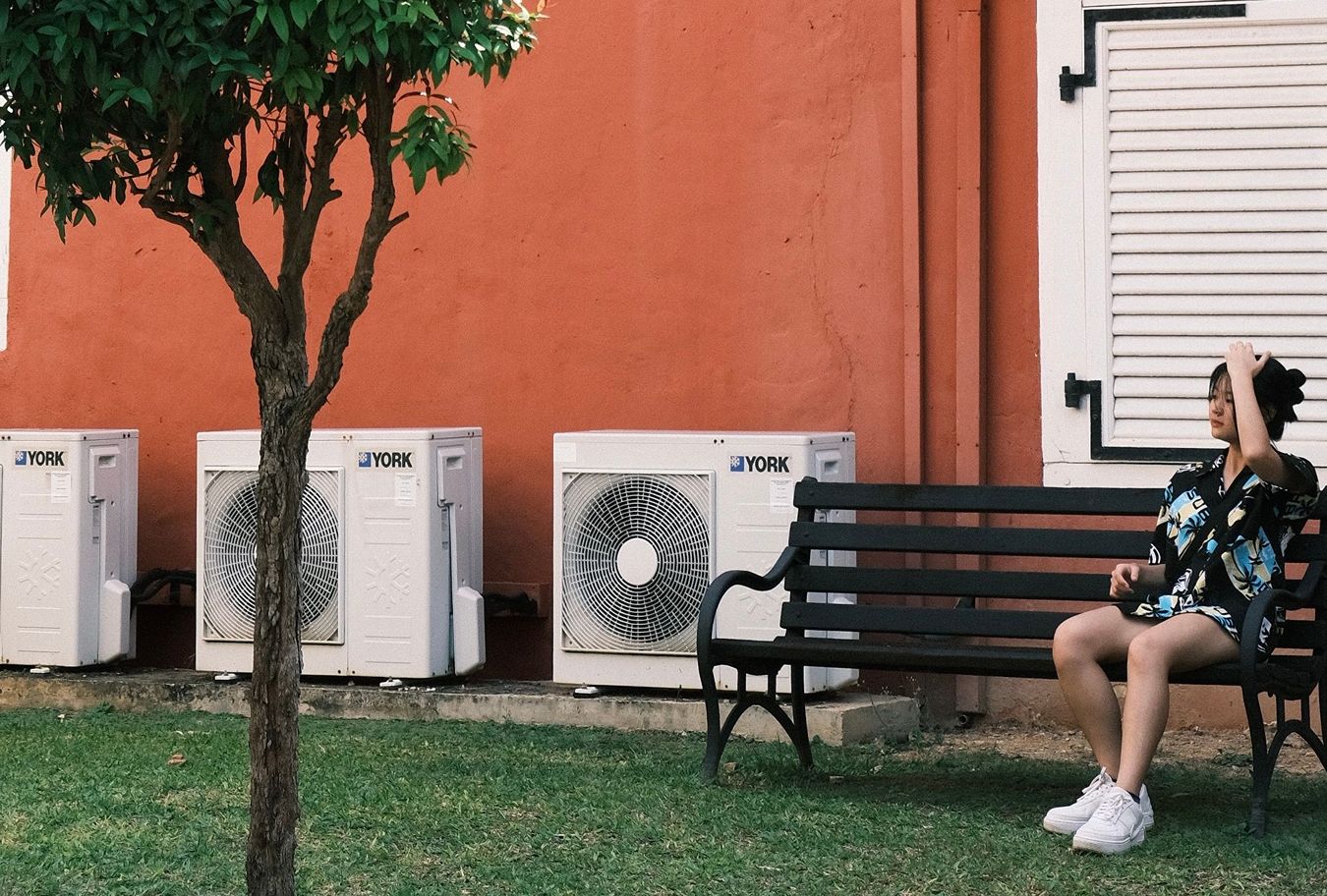 A woman sitting on a bench near AC units outside a building.