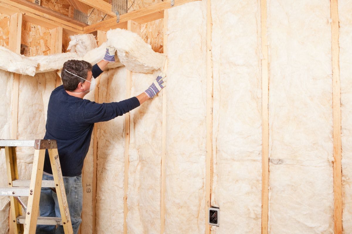 A person on a letter installs insulation between wall studs.