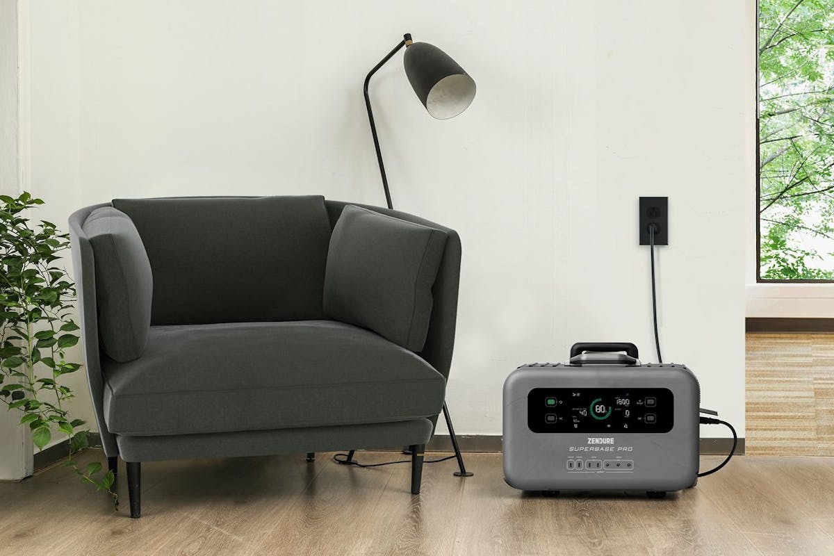 A portable power station plugged into the wall by an arm chair and a lamp.