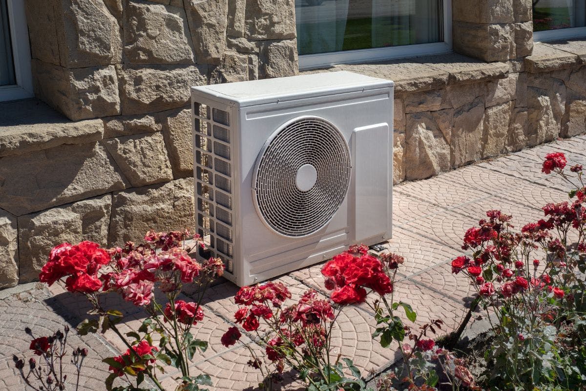 A heat pump near a brick building surrounded by red flowers.