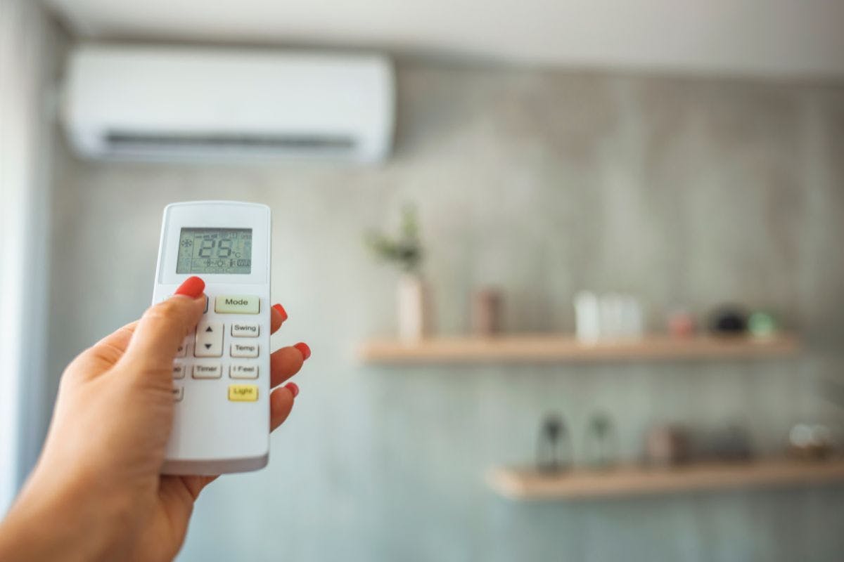 A remote held by a hand with painted fingernails pointing at a mini-split air conditioning or heat pump unit mounted on the wall.