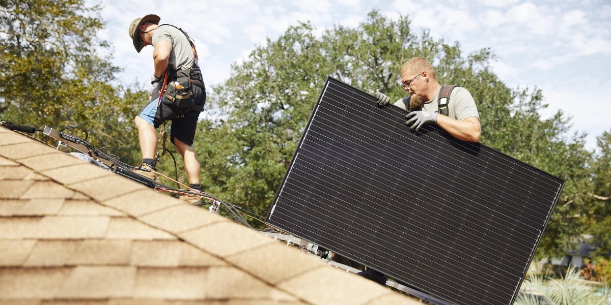 Two solar installers move a black solar panel on a roof.