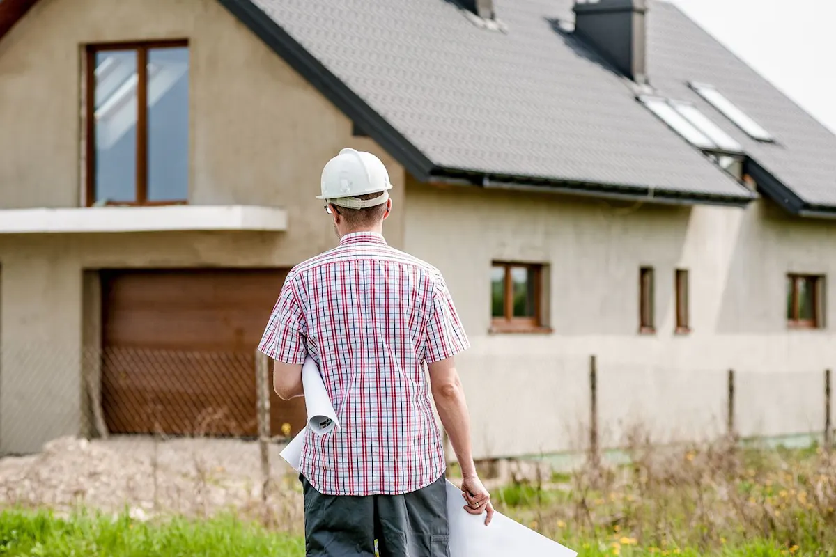 A worker with plans in hand walks toward a modern house.