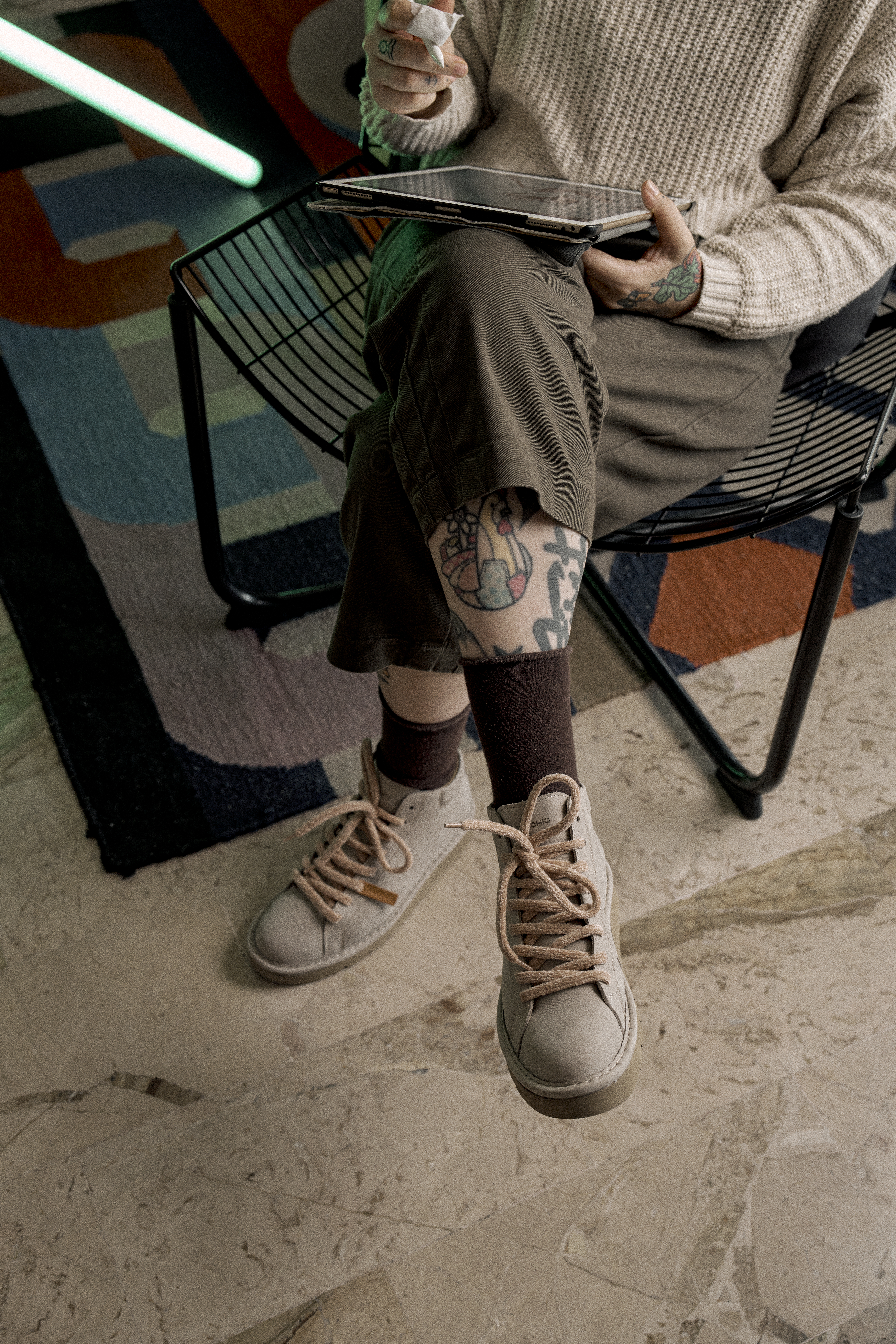 A woman is sitting crossed-legged on a chair wearing a pair of dove-grey P007 ankle boots. Above her socks we can see her tattoos.