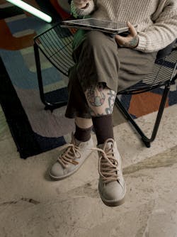 A woman is sitting crossed-legged on a chair wearing a pair of dove-grey P007 ankle boots. Above her socks we can see her tattoos.