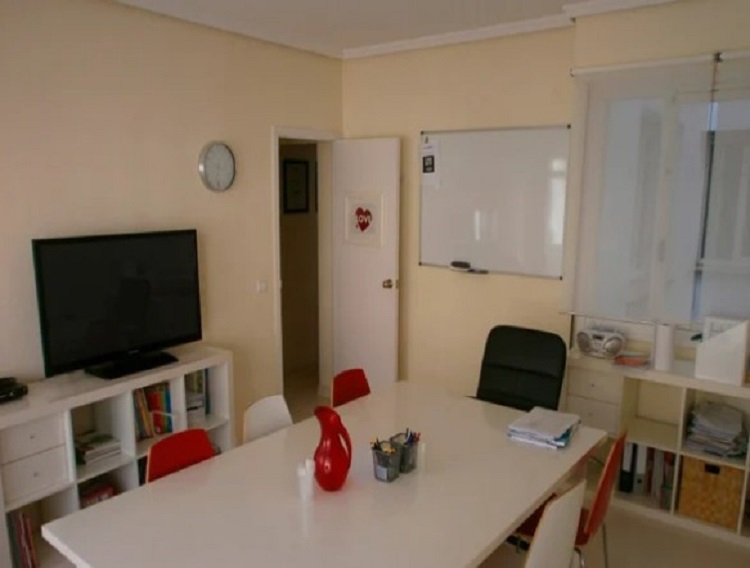 Salon de clases con pizarra, mesa, un televisor y sillas rojas y blancas | Salones del curso de ingl&eacute;s Think English