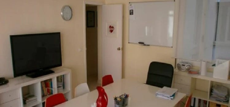 Salon de clases con pizarra, mesa, un televisor y sillas rojas y blancas | Salones del curso de inglés Think English
