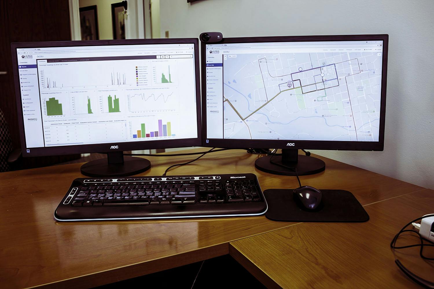 Dual computer monitors displaying Passio OpsView™ with real-time transit maps and performance dashboards, used for tracking vehicle activity, system metrics, and operational alerts.