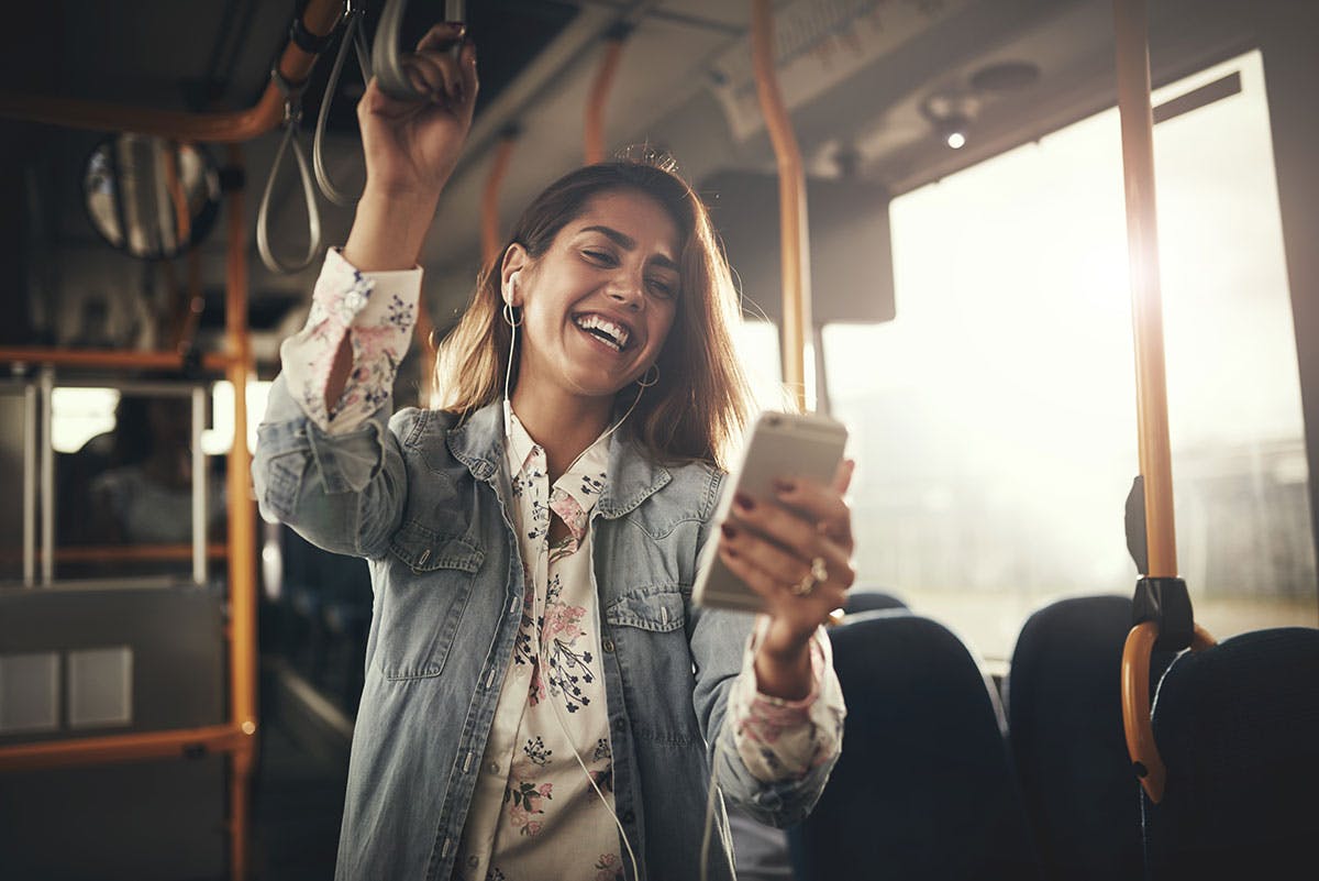 Smiling woman riding a bus, holding onto a handrail and looking at her smartphone while listening to music with earbuds, with sunlight streaming through the window.