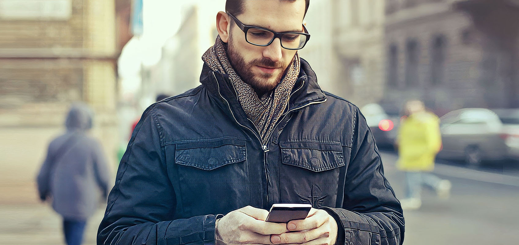 Man standing on a city sidewalk using a smartphone, representing a transit passenger engaging with the Passio GO™ app to plan and track a bus trip in real time.