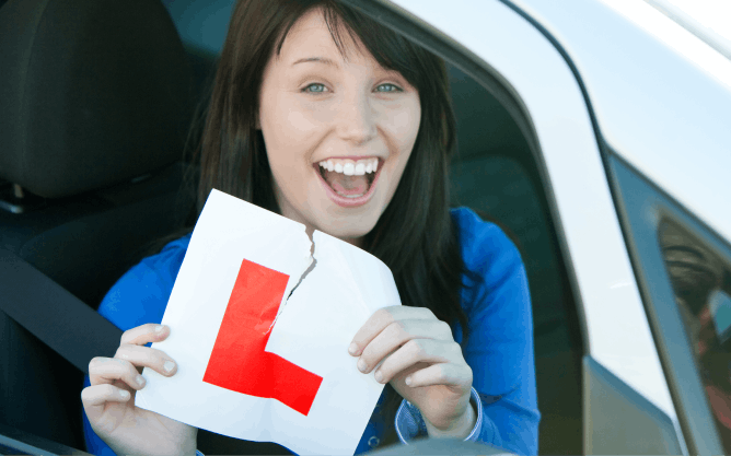 Photograph of a lady sat in a car. She is holding a learner driver L plate out of the window and smiling as she rips it, suggesting she has just passed her driving test.