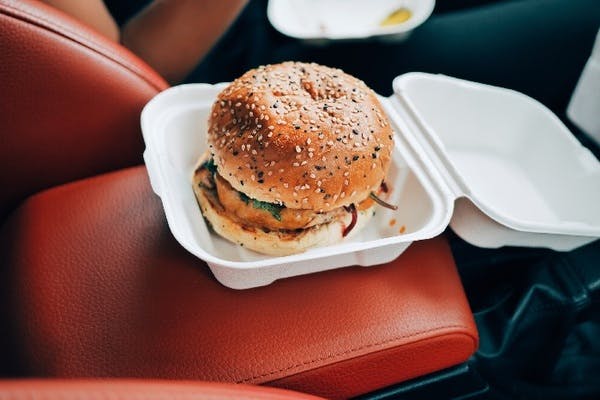 A large burger with a seeded bun in a takeaway box on a leather car seat
