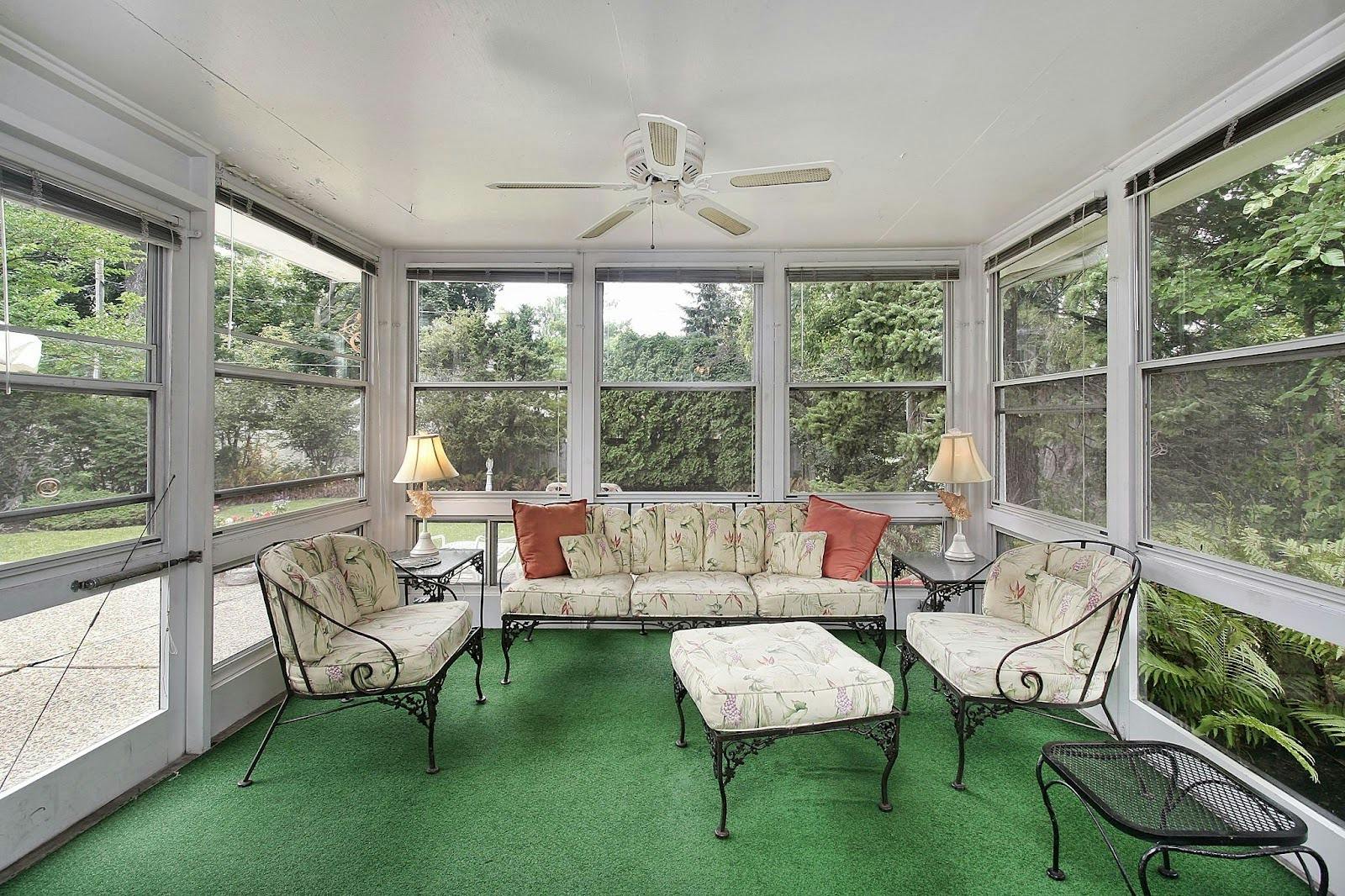 Bright enclosed patio space designed as a three-season room with screened windows, a white ceiling fan, floral cushioned seating, and green carpet flooring surrounded by trees outside.