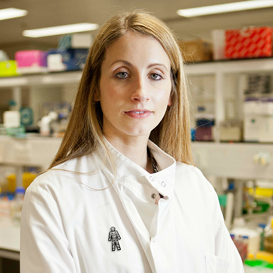 Female researcher with long blonde hair, wearing lab coat and Prostate Cancer UK badge inside a lab.