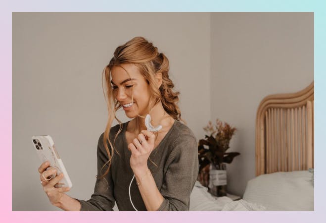 A girl in her bedroom, looking at her phone while holding the Pearlii Home Teeth Whitening Kit mouth tray