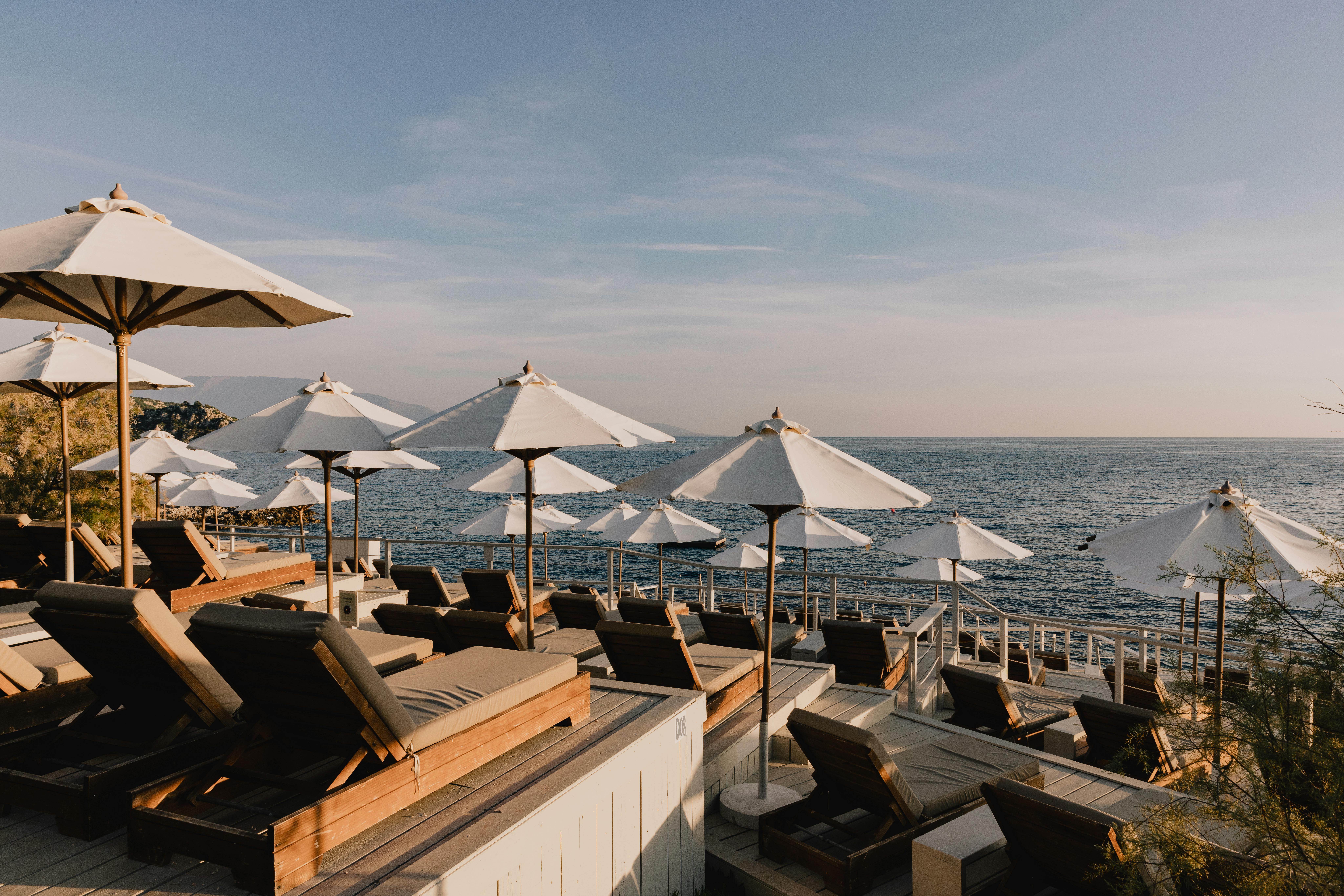 A luxurious beachfront terrace featuring multiple white canvas umbrellas and dark wooden loungers arranged in rows. The scene overlooks a calm ocean at dusk with a soft blue-pink sky. The tiered deck design creates an elegant outdoor lounge space with tropical vegetation visible at the edges. The setting appears to be at an upscale resort or beach club