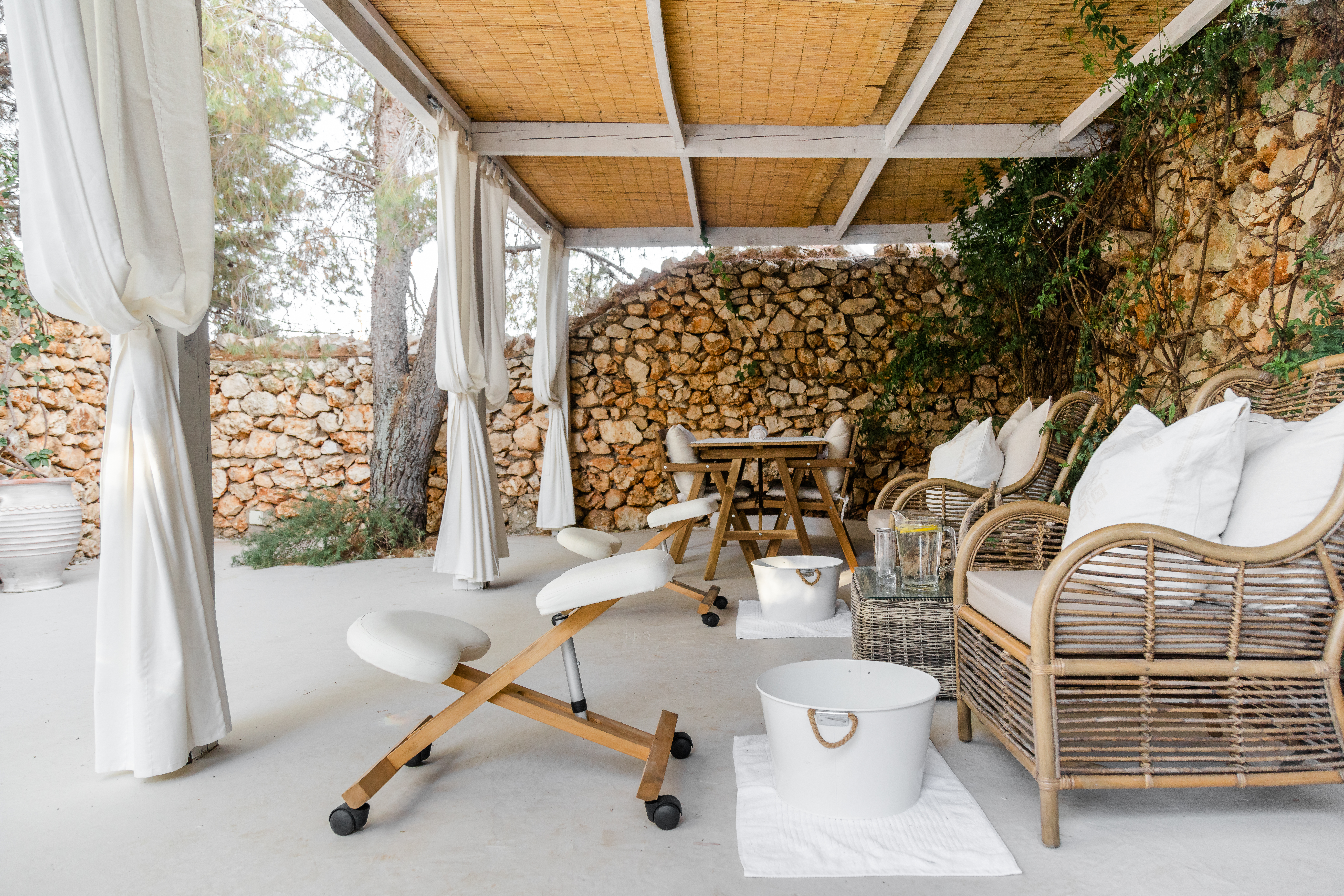 A serene outdoor spa relaxation area featuring a covered terrace with natural bamboo reed ceiling and white wooden beams. The space includes a white cushioned massage chair with wooden frame on wheels, rattan lounge chairs with plush white pillows, and a white foot bath basin with rope handles. The backdrop showcases rustic stone walls with climbing vines and Mediterranean vegetation. A small wooden side table sits between seating areas, creating an intimate wellness retreat atmosphere.