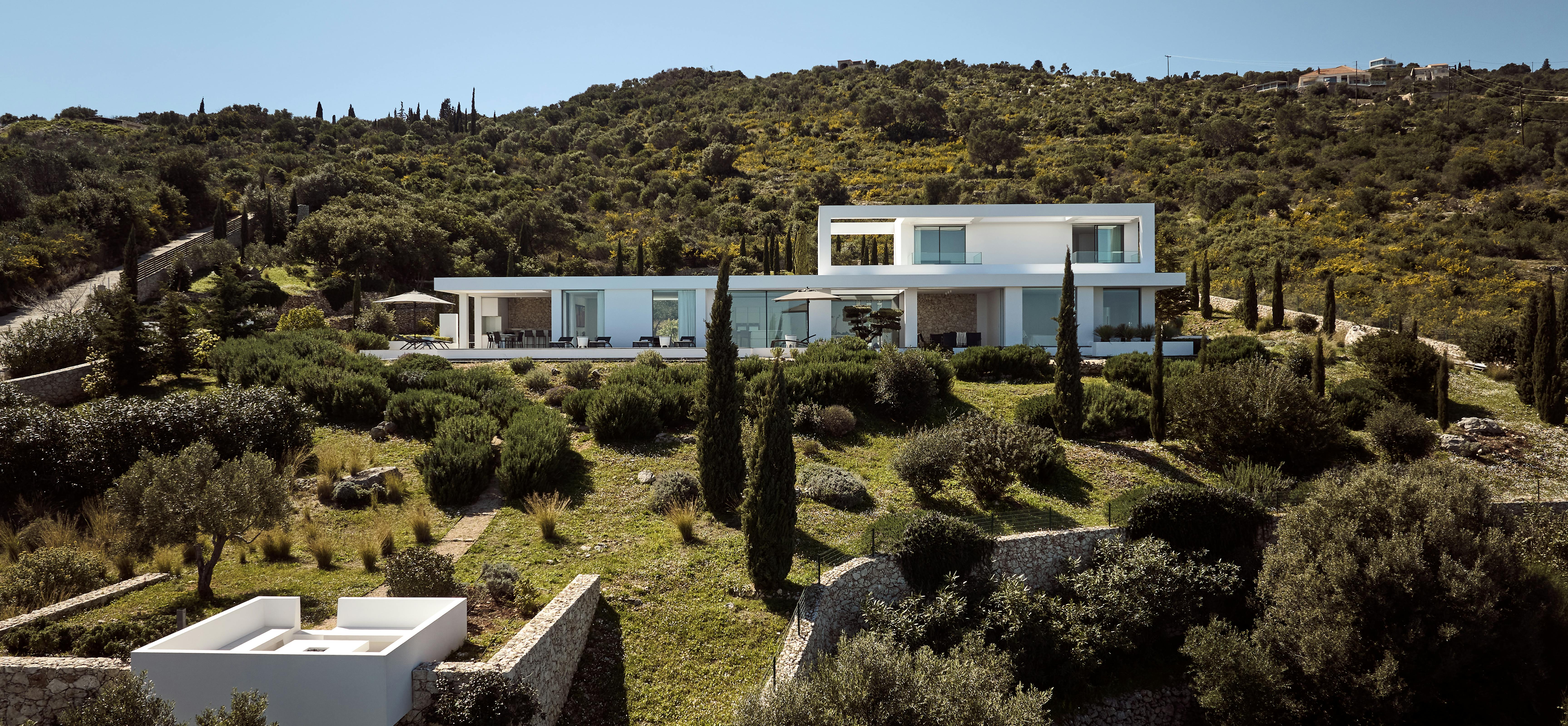 Aerial view of contemporary white villa with glass walls nestled among olive trees and cypress on Mediterranean hillside with small plunge pool.