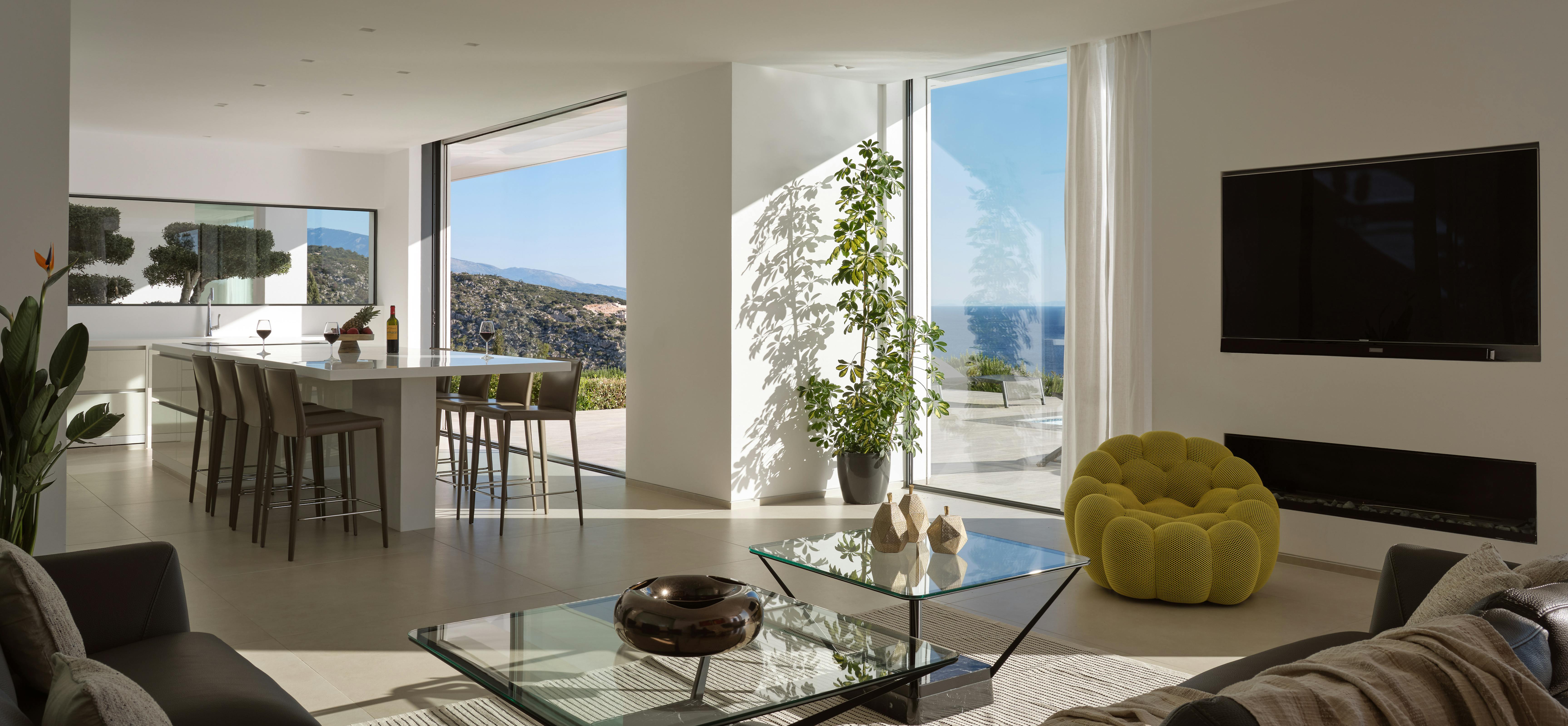 Open-plan living space with grey sectionals, glass coffee table, chartreuse bubble chair, and sliding doors to hillside dining terrace with sea beyond.