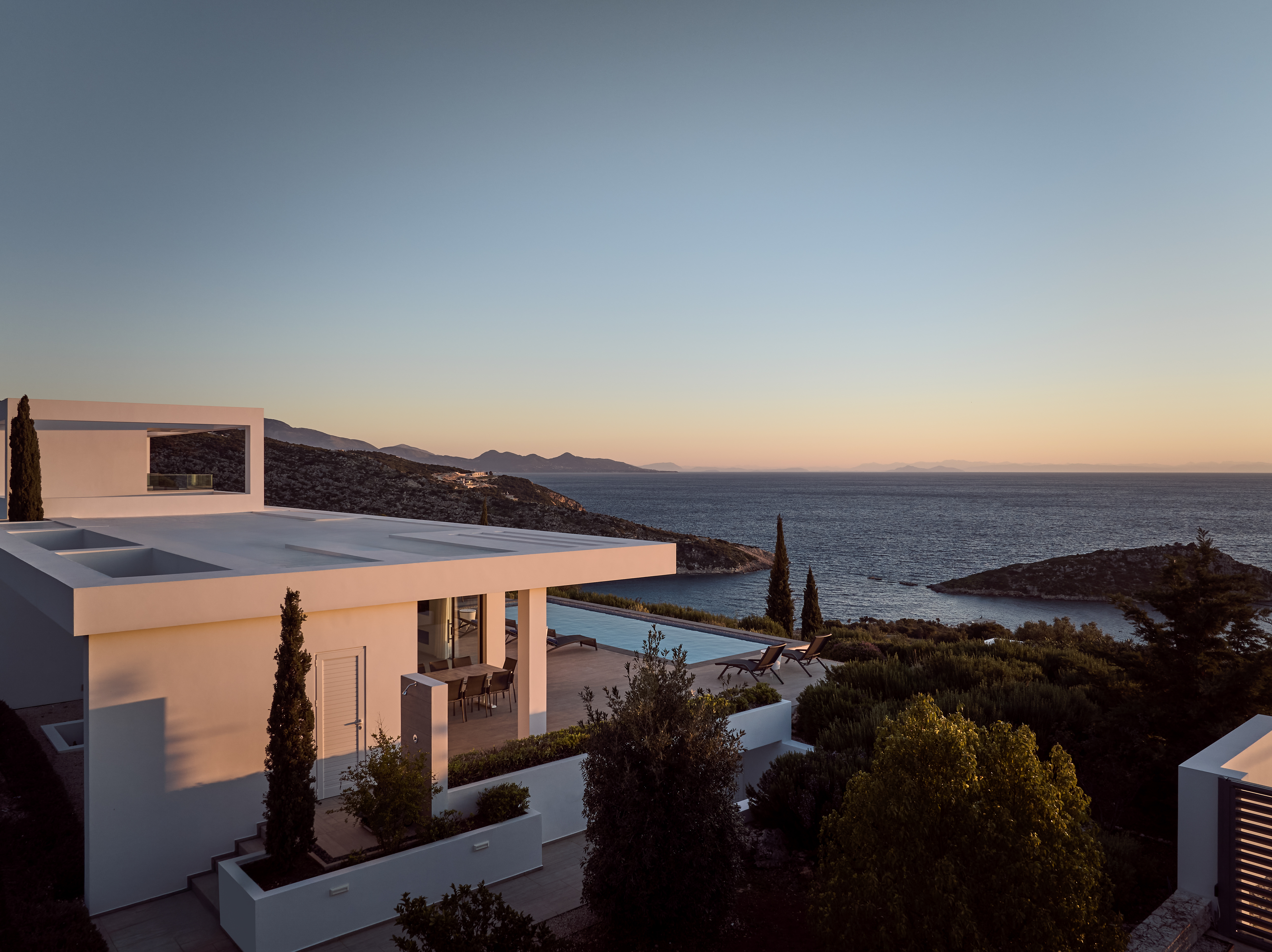 Modern white villa at golden hour with stacked geometric volumes, covered terraces, infinity pool, and sweeping sea views toward rocky cove.