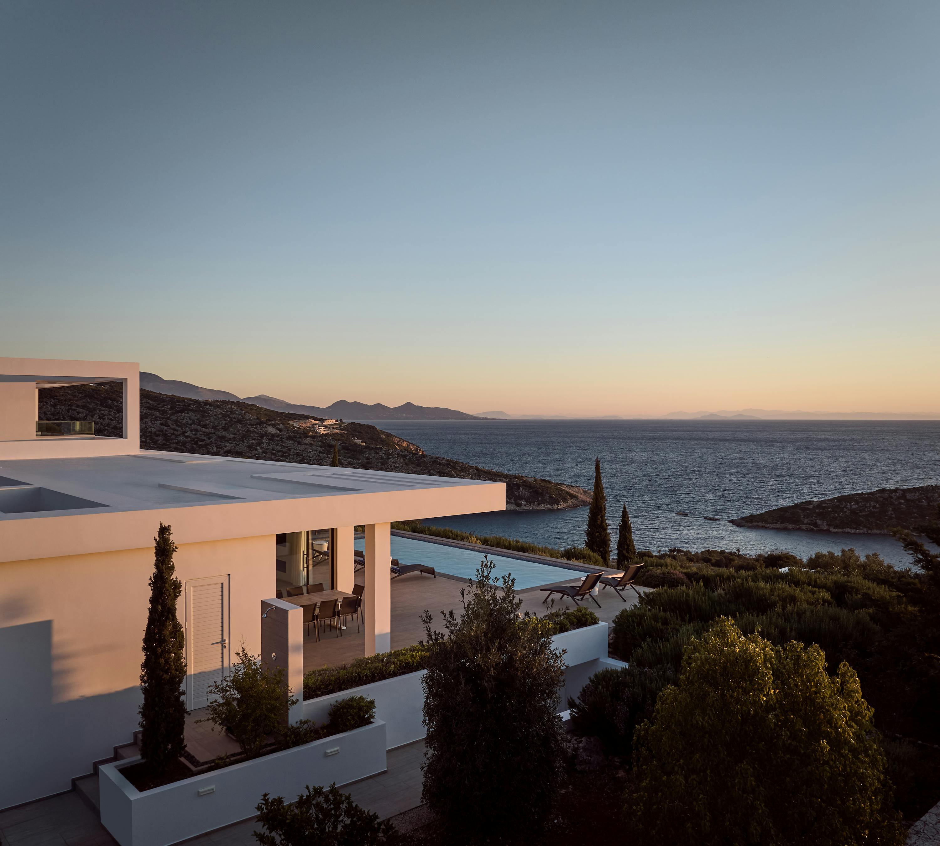 Modern white villa at golden hour with stacked geometric volumes, covered terraces, infinity pool, and sweeping sea views toward rocky cove.