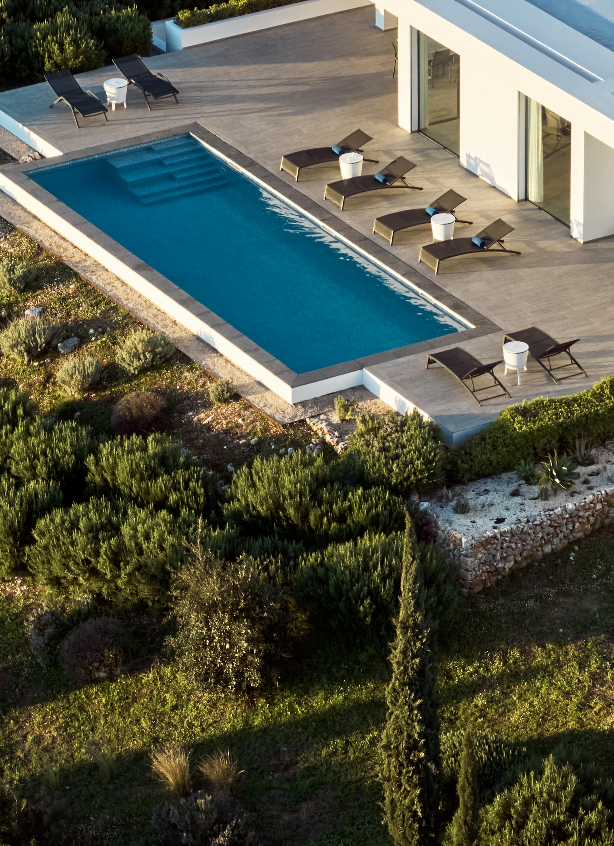 Aerial view of rectangular infinity pool with dark loungers, white side tables, and wood deck surrounded by Mediterranean scrubland.