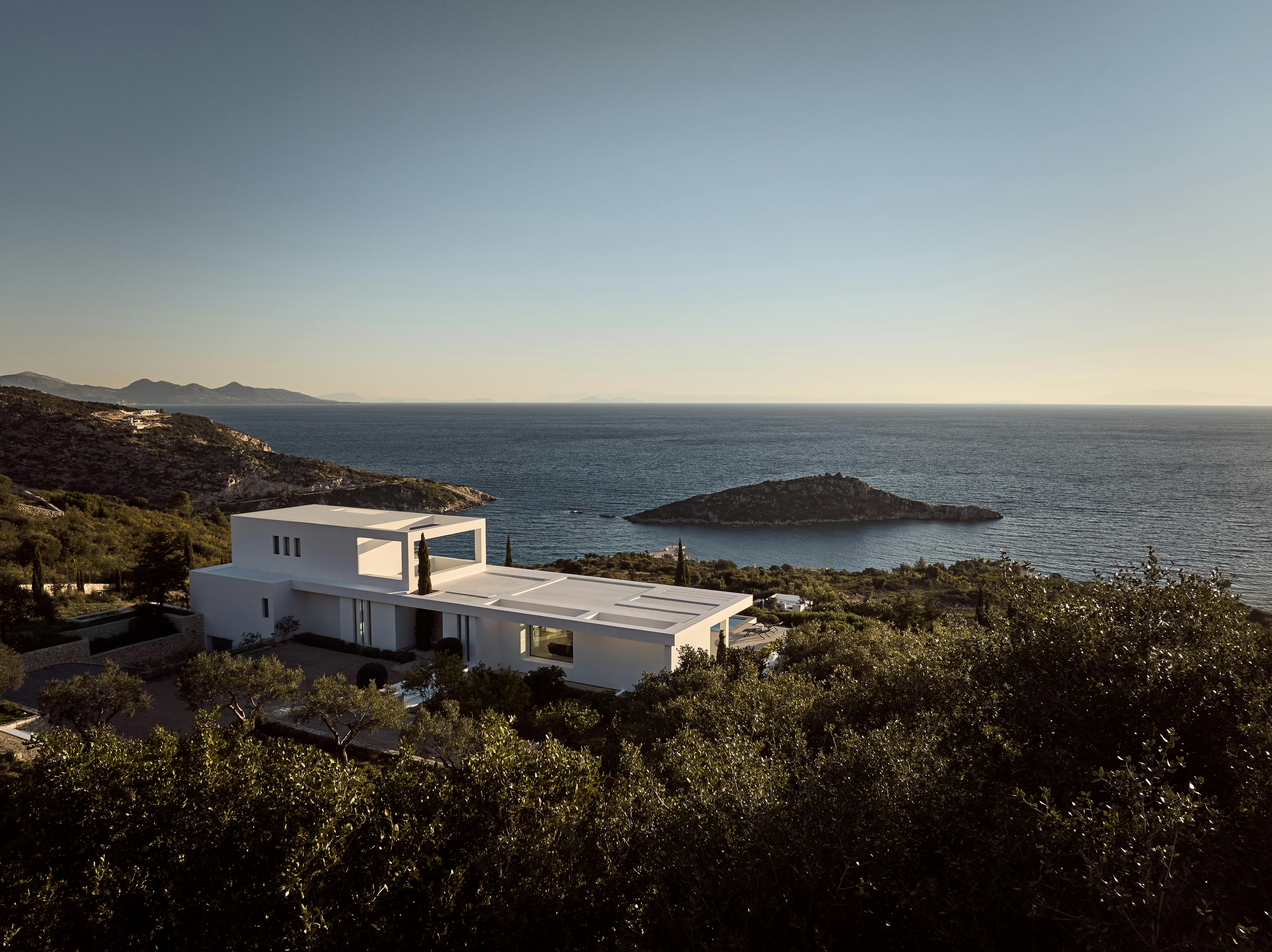 White modernist villa at dusk perched on hillside overlooking calm bay with small island, surrounded by dense Mediterranean vegetation and cypress.