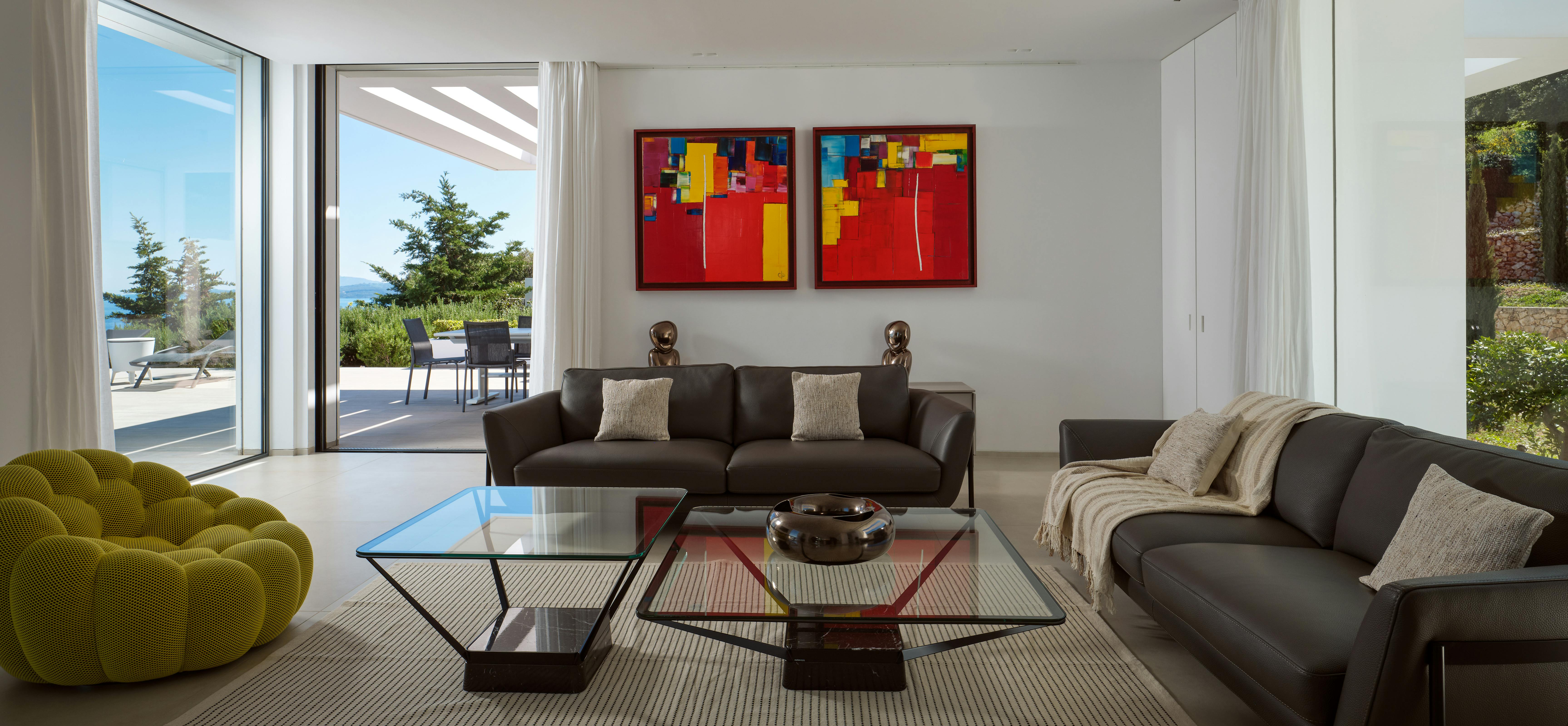 Modern living room with grey sofas, vibrant abstract red and yellow artwork, glass coffee table, and terrace doors framing sea views.