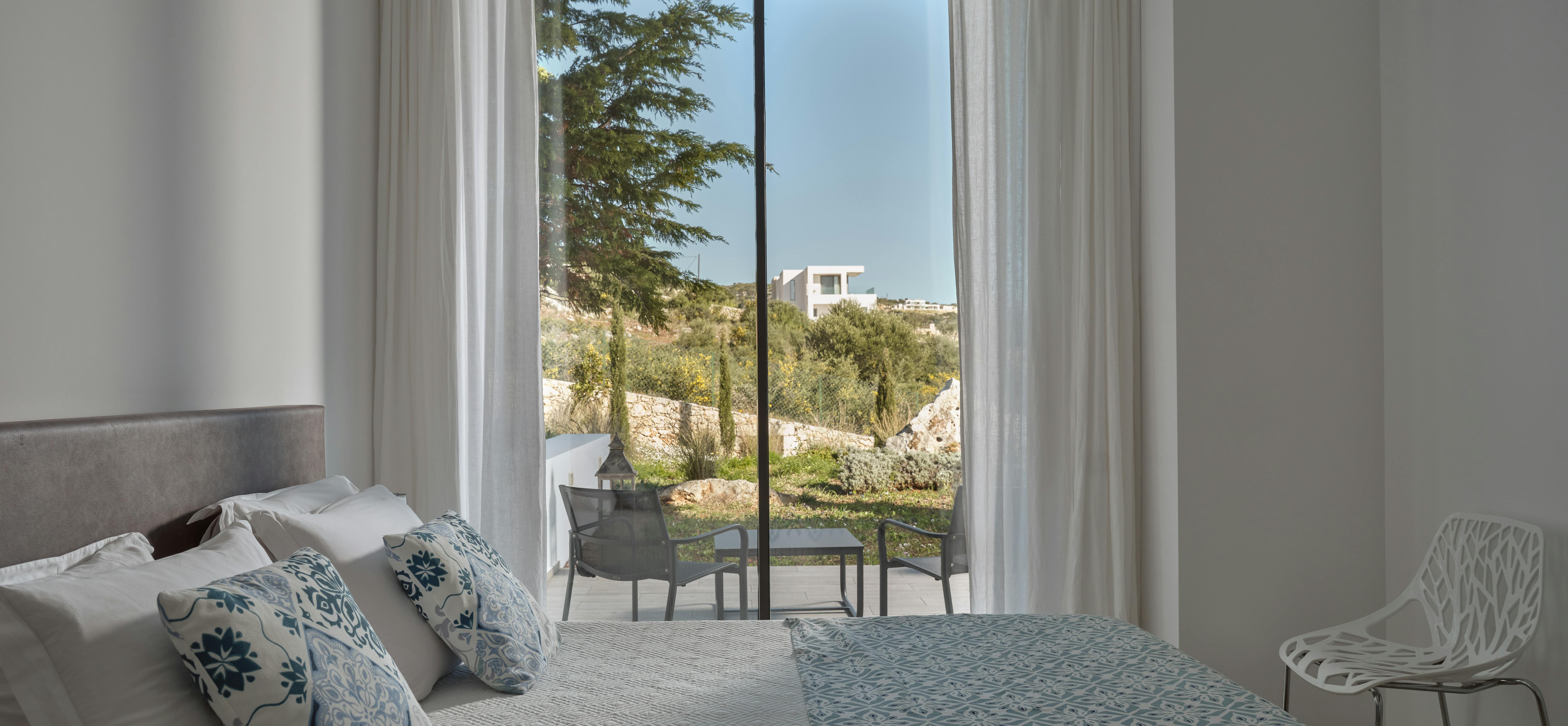 Serene bedroom with grey headboard, blue patterned linens, white chair, and sliding glass doors opening to private terrace overlooking olive groves.