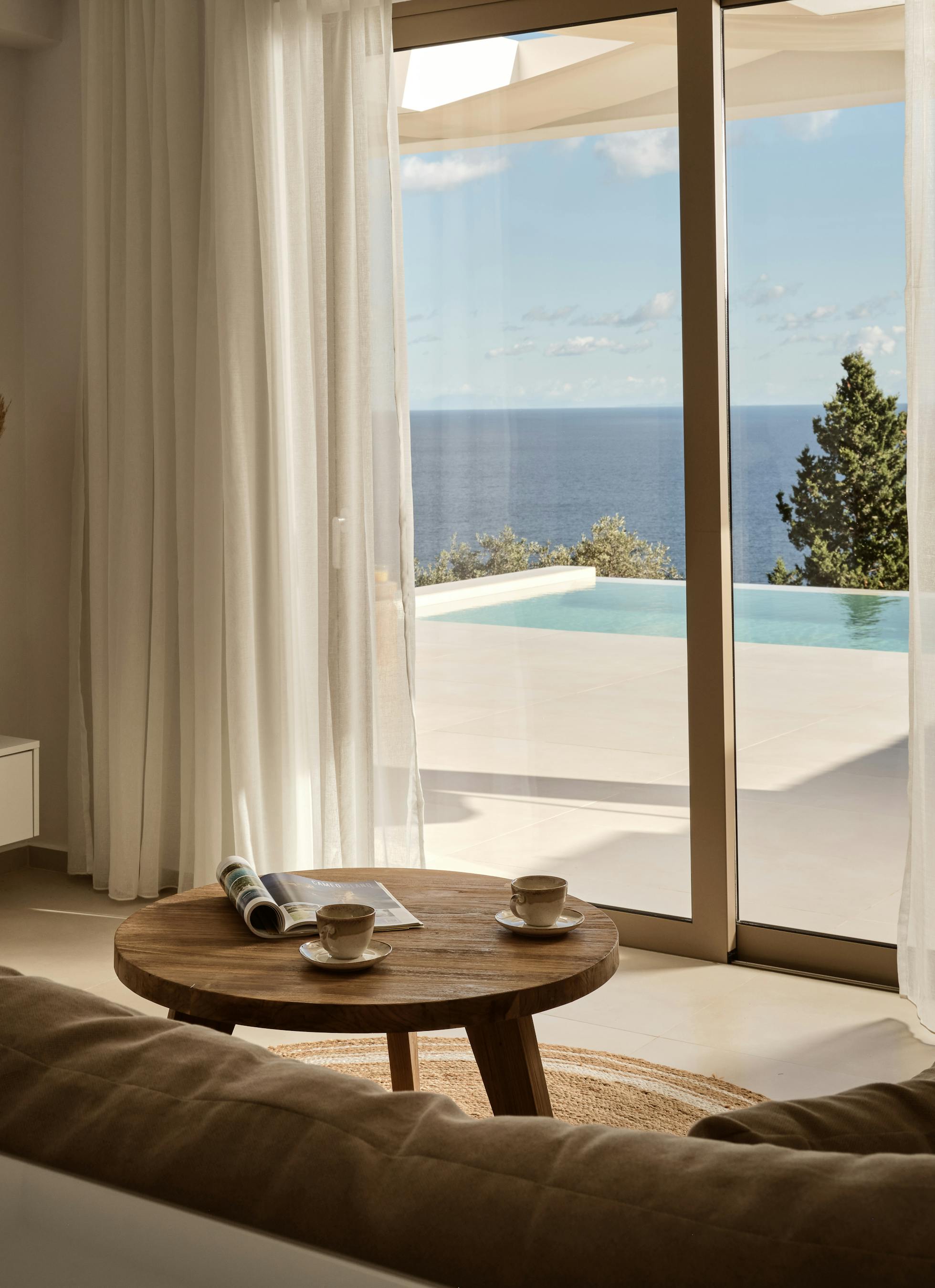 Contemporary living space with woven wall baskets, wood furniture, and floor-to-ceiling windows overlooking pool and sea.