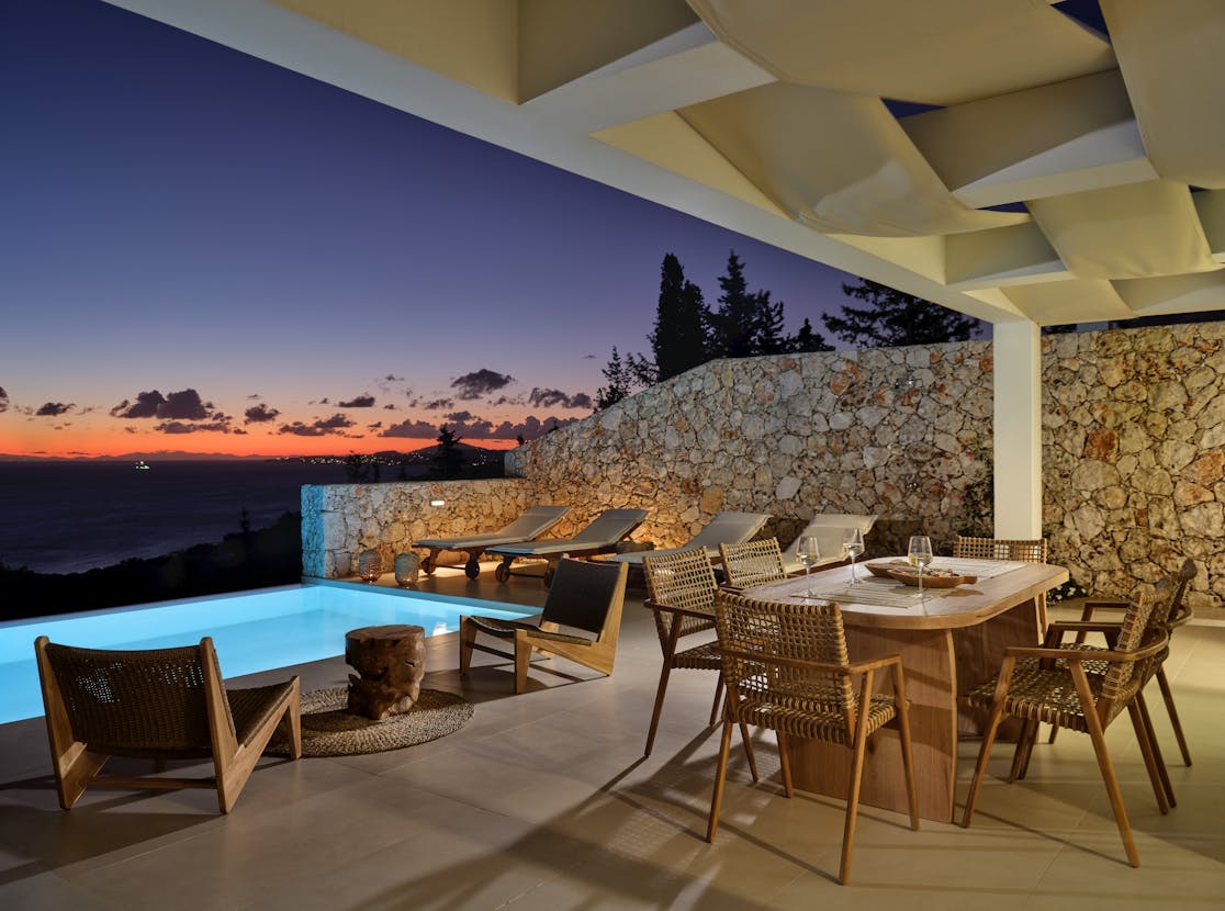 Evening terrace scene with illuminated pool, dining area, and dramatic purple-orange sunset sky.