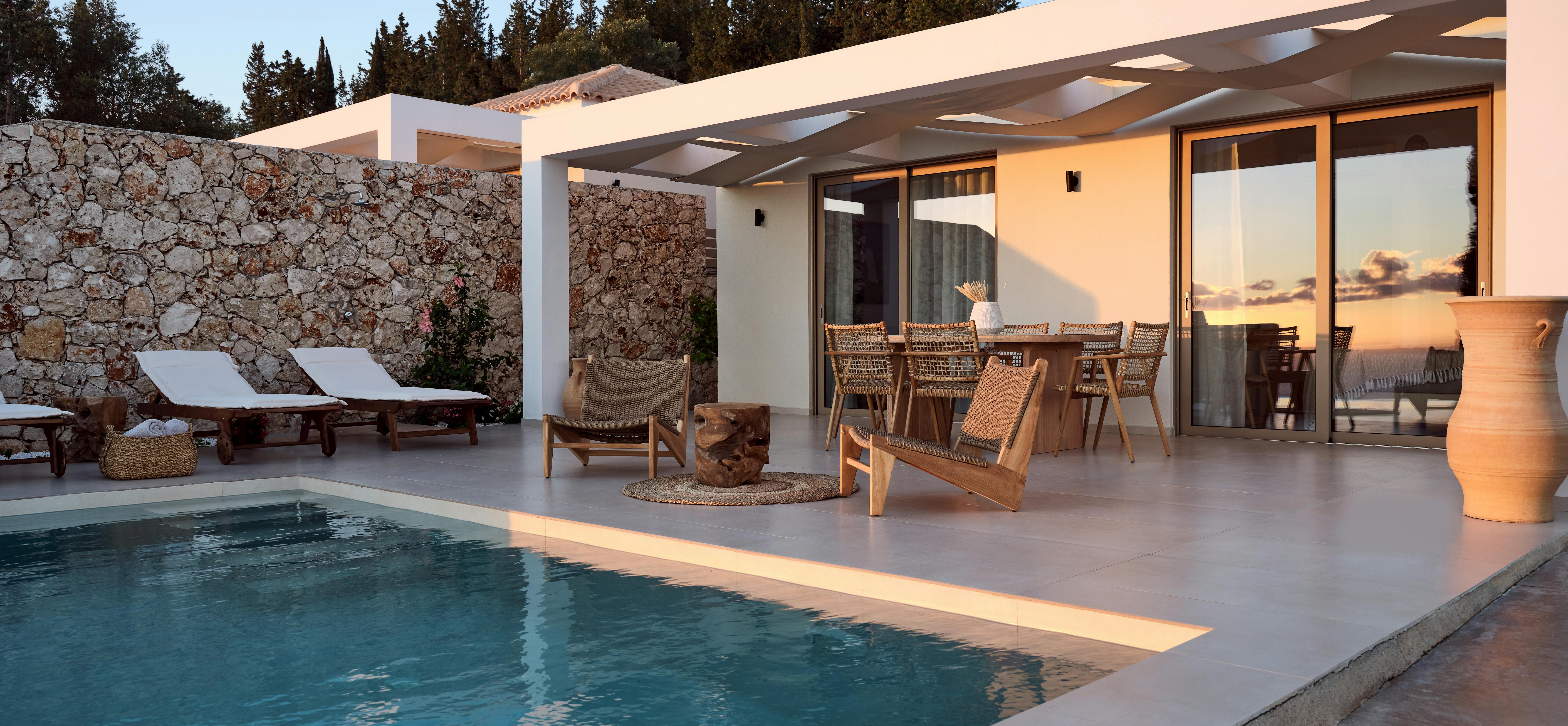 Villa pool terrace at dusk with illuminated stone walls and woven furniture facing dramatic sunset.