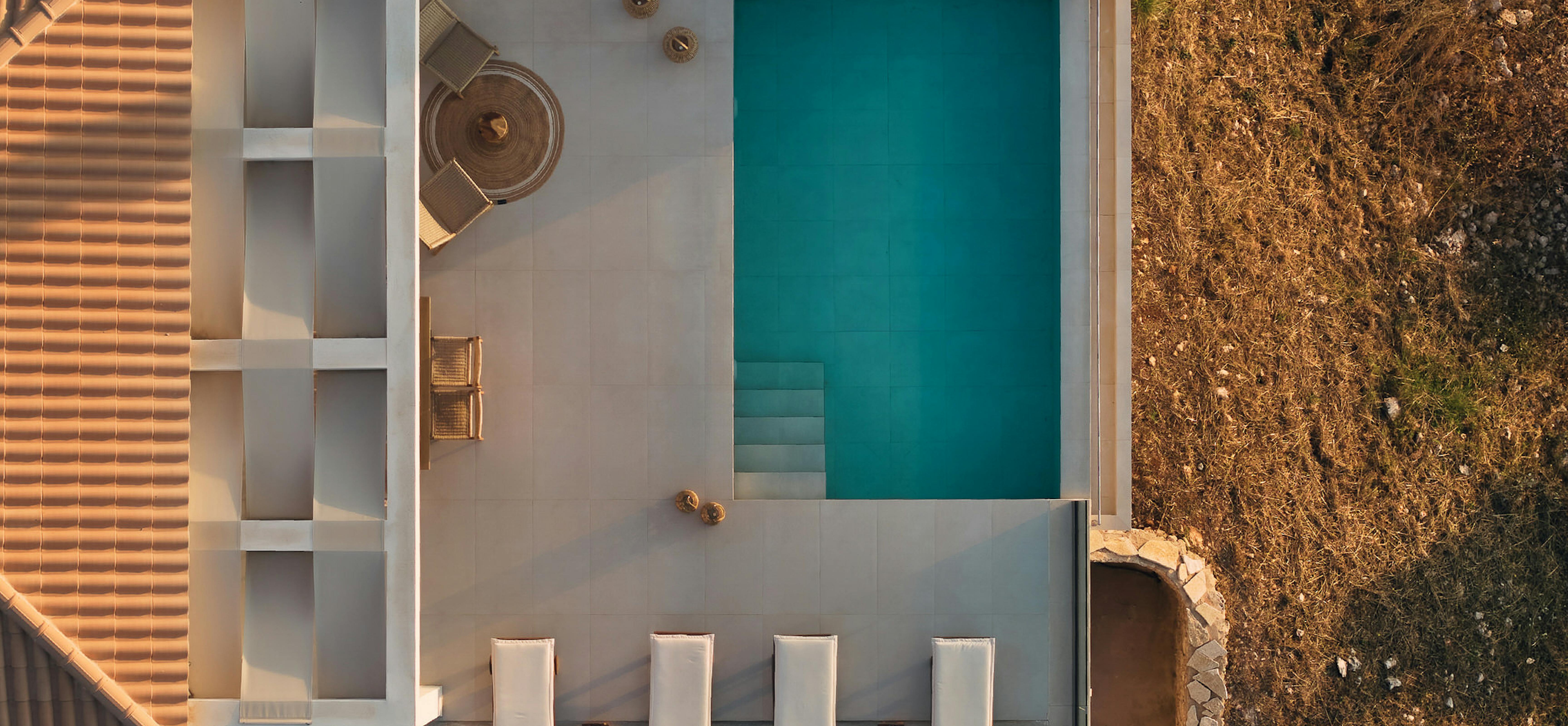 Bird's-eye view of villa courtyard showing terracotta roofs, rectangular pool, sun beds, circular fire pit, and arid hillside.