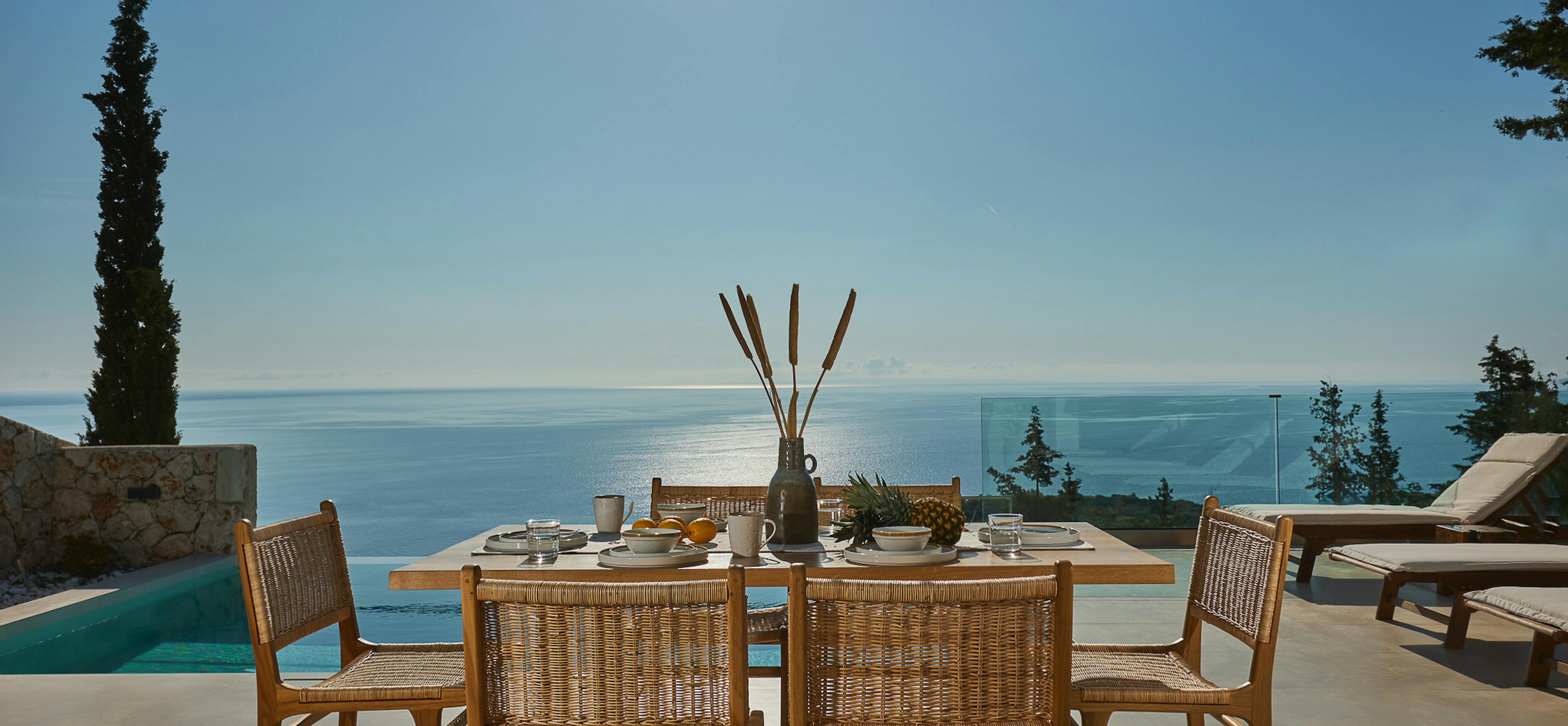 Outdoor breakfast table with woven chairs set for morning meal on infinity pool terrace overlooking shimmering sea and cypress trees.