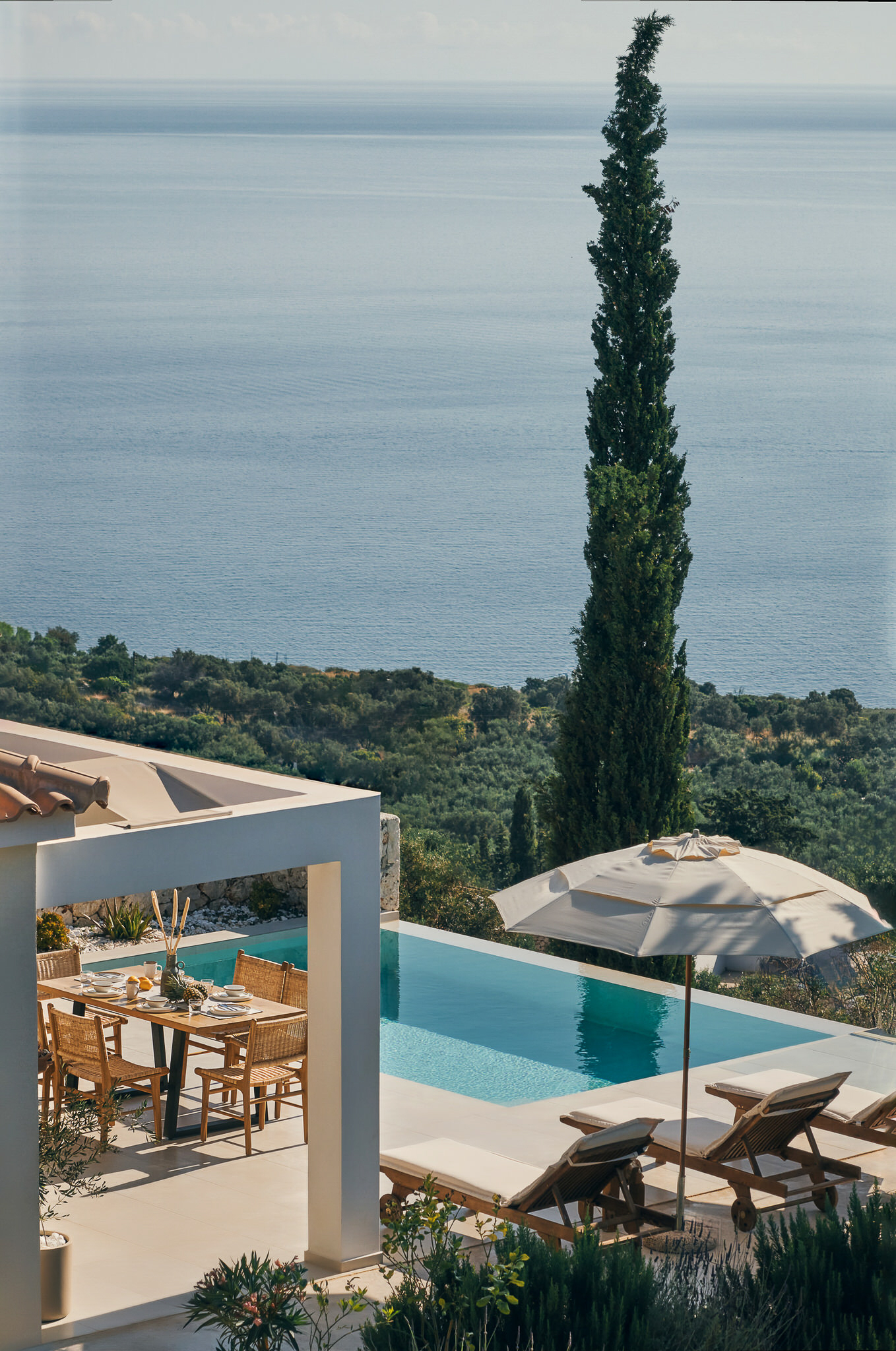 Elevated view of multi-level villa terraces with infinity pool, white umbrella, loungers, tall cypress, and coastal vista beyond olive groves.