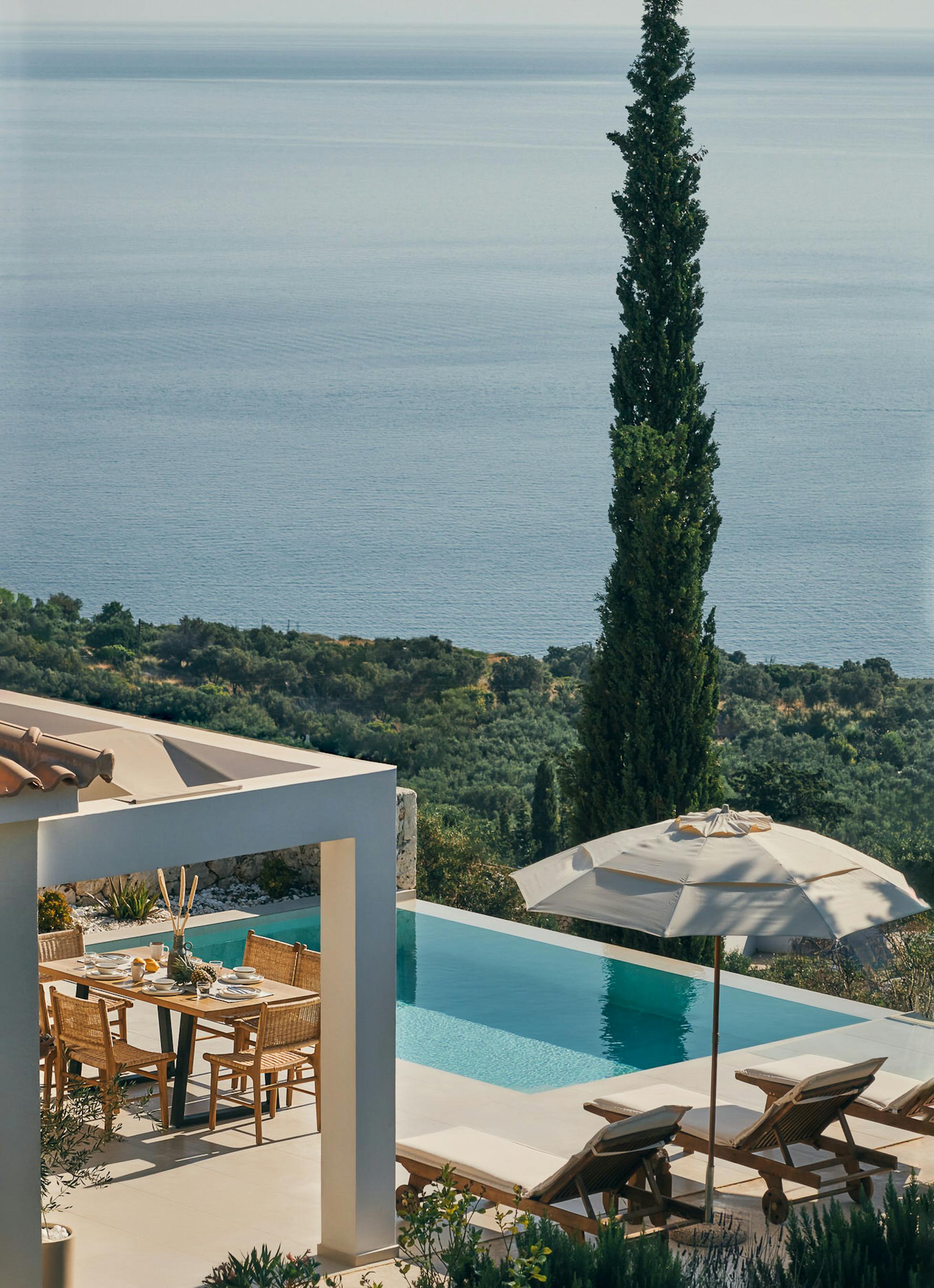 Elevated view of multi-level villa terraces with infinity pool, white umbrella, loungers, tall cypress, and coastal vista beyond olive groves.