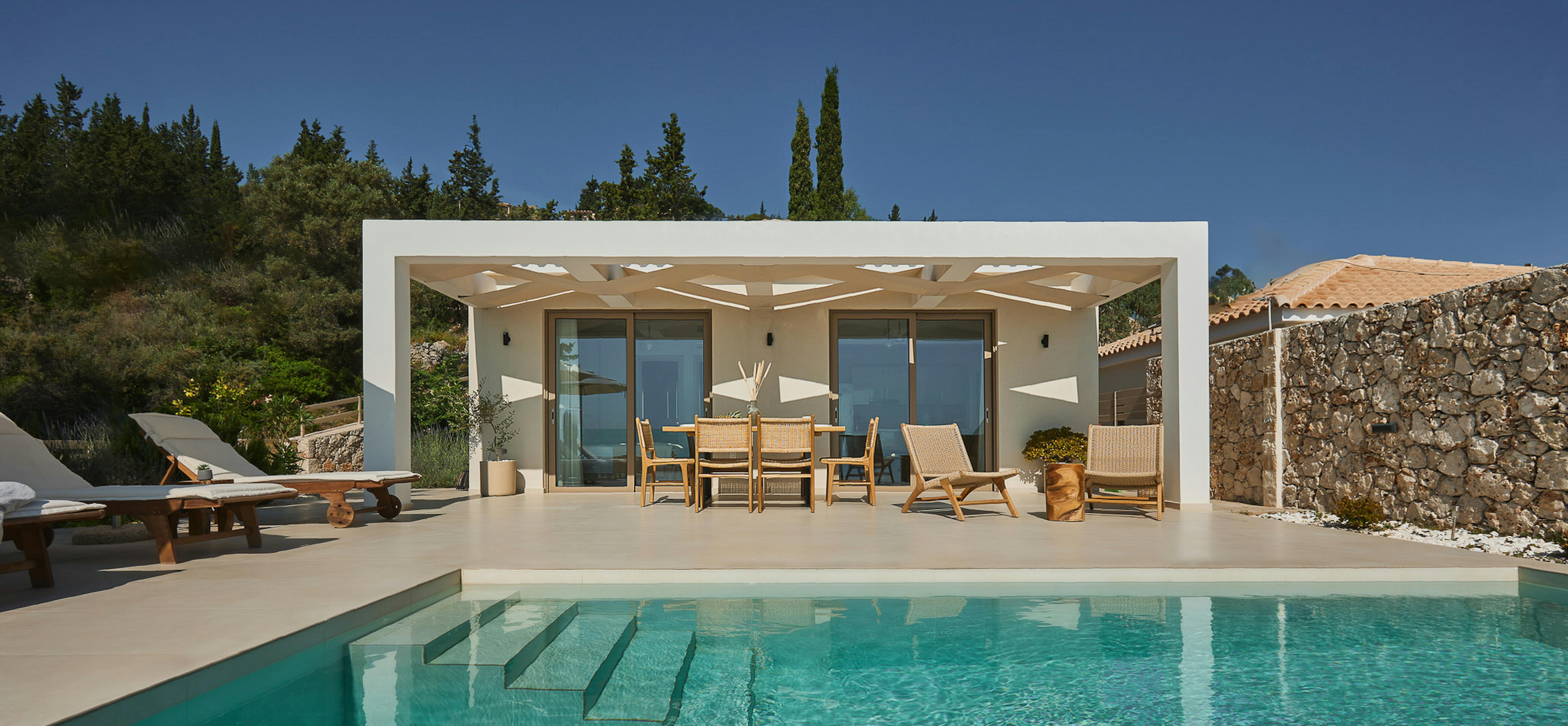 Modern pool pavilion with white pergola, glass doors, woven dining furniture, loungers with submerged steps, backed by stone wall and cypress trees.