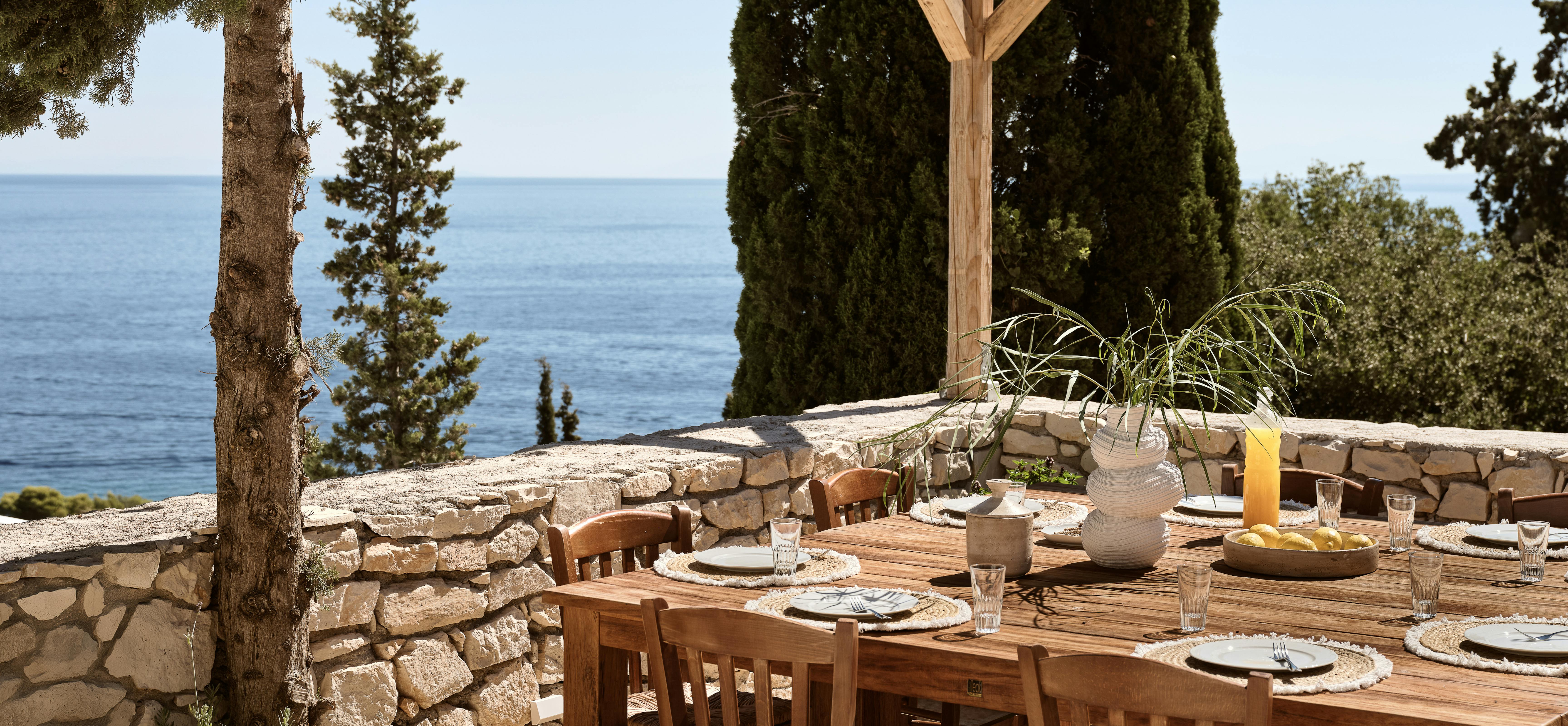 An outdoor dining table set with wooden chairs sits under string lights on a stone-walled terrace overlooking the sea, with cypress trees framing the view.