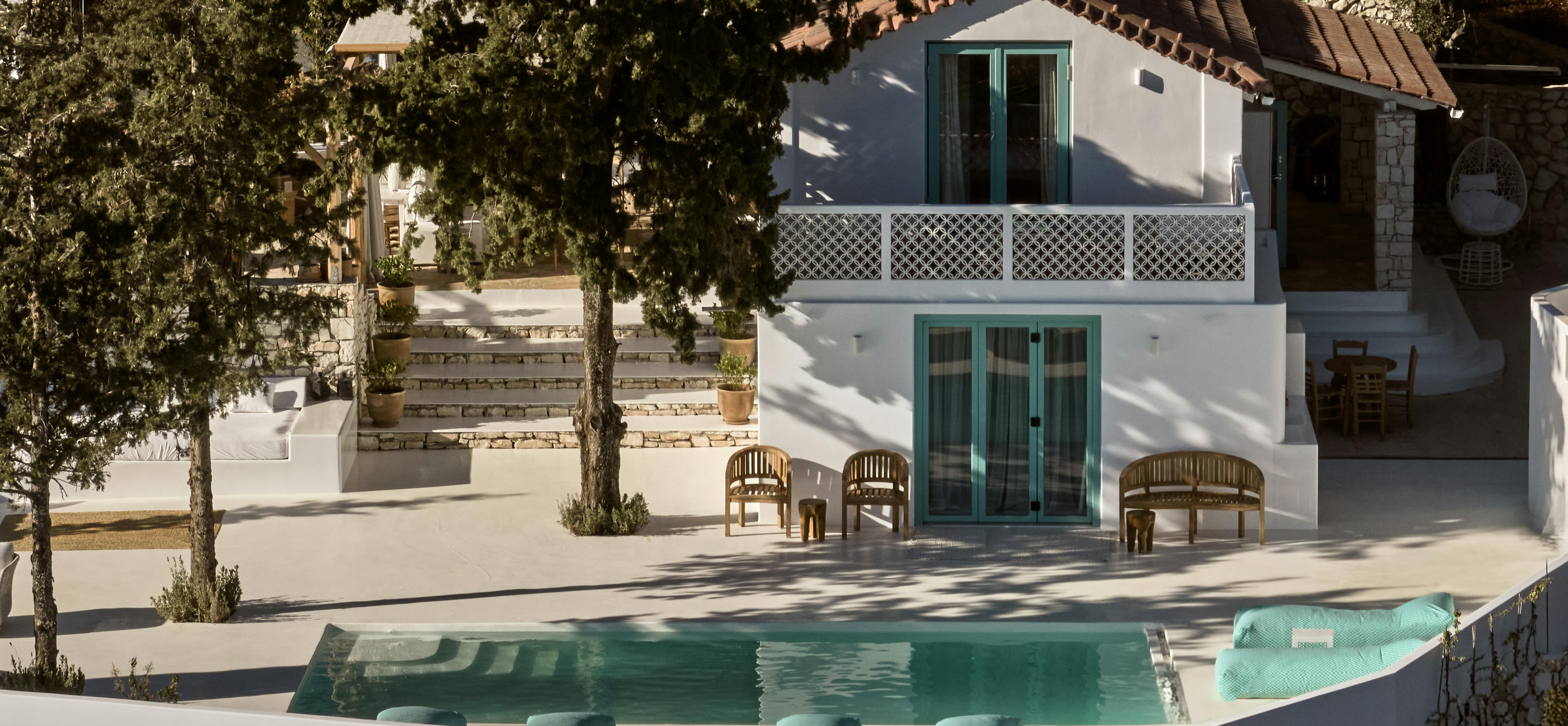A white Mediterranean villa with turquoise-trimmed windows surrounds a turquoise pool, with wooden furniture arranged on white terraces amid olive trees.