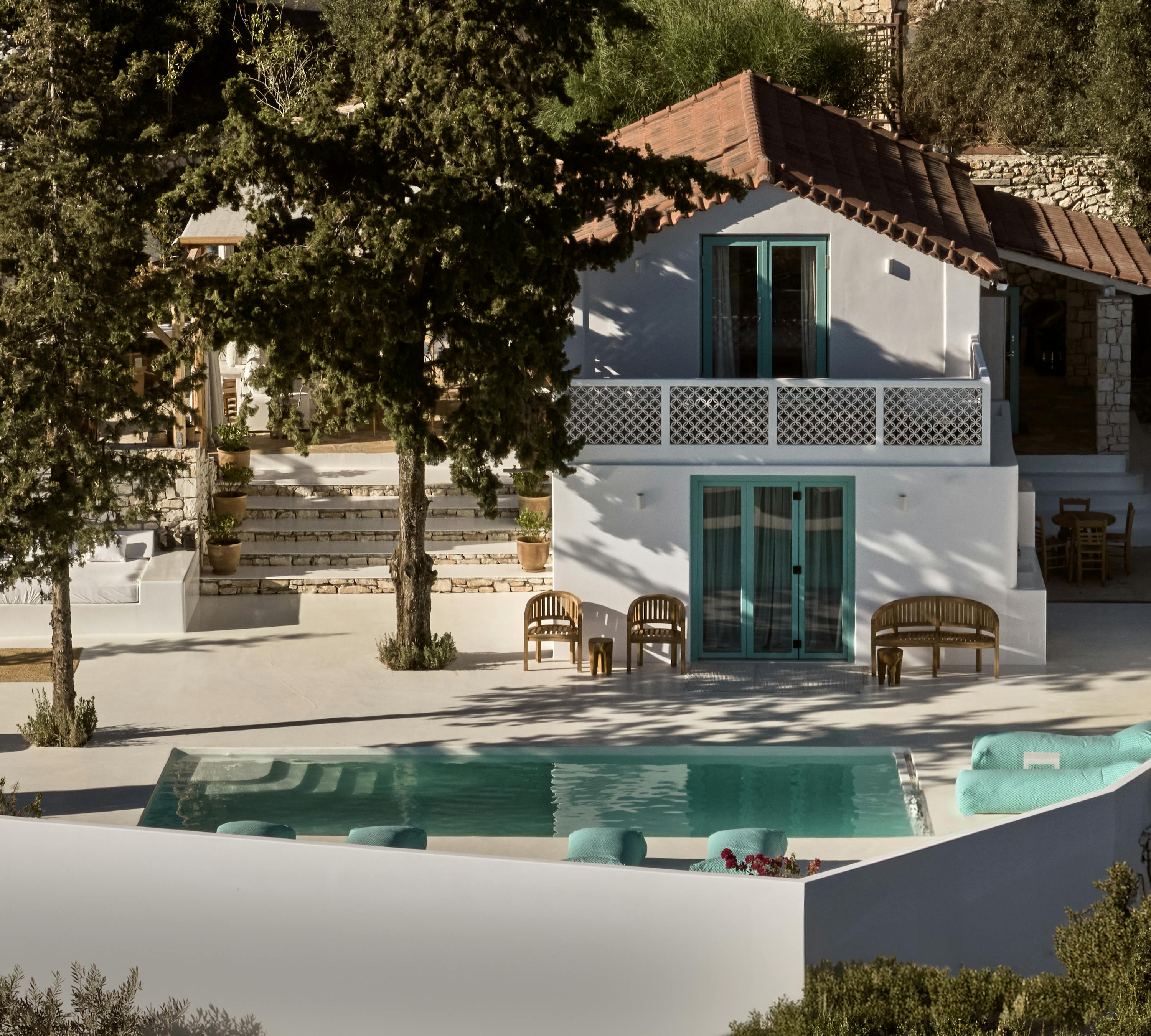 A white Mediterranean villa with turquoise-trimmed windows surrounds a turquoise pool, with wooden furniture arranged on white terraces amid olive trees.
