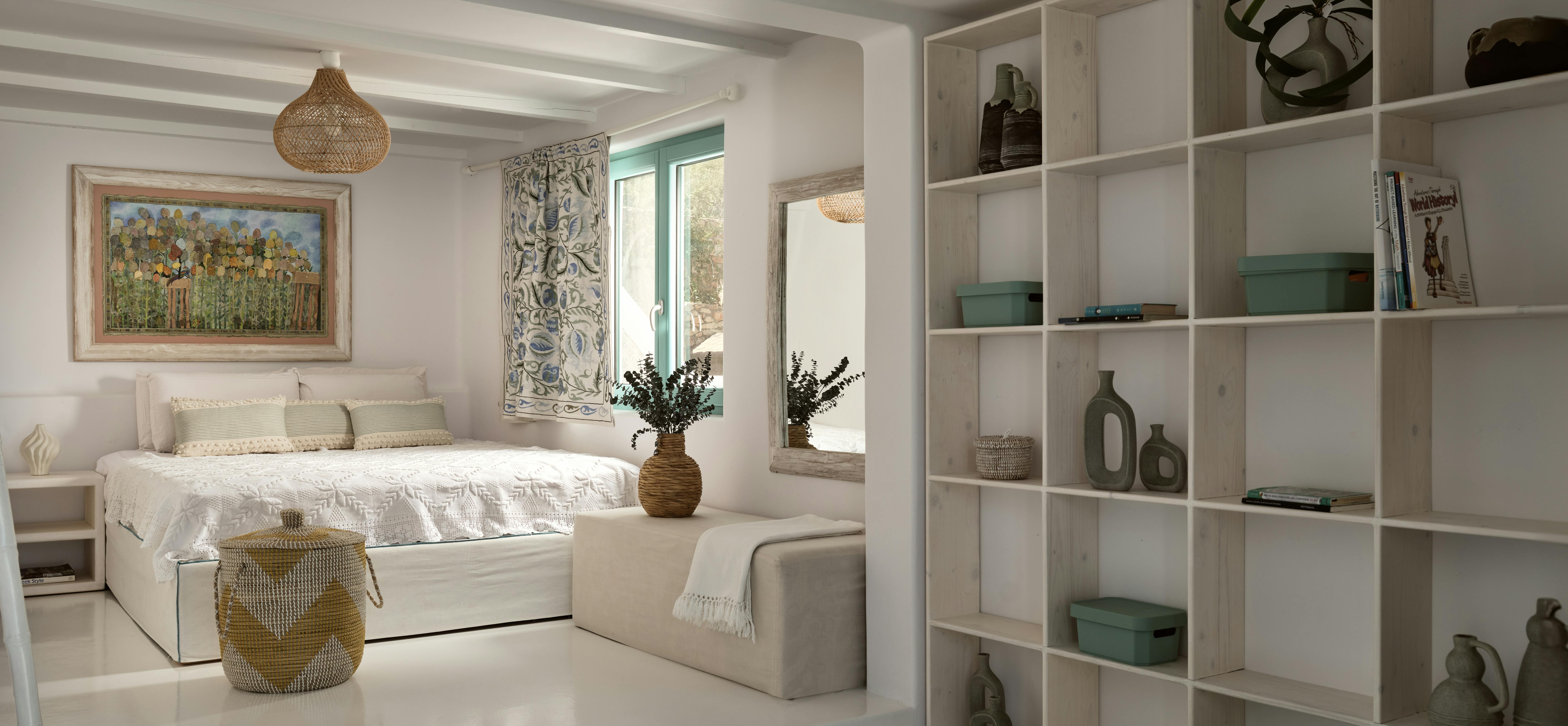 A bright white bedroom suite with exposed beam ceilings includes a platform bed, colourful artwork, built-in shelving, and woven pendant lighting.
