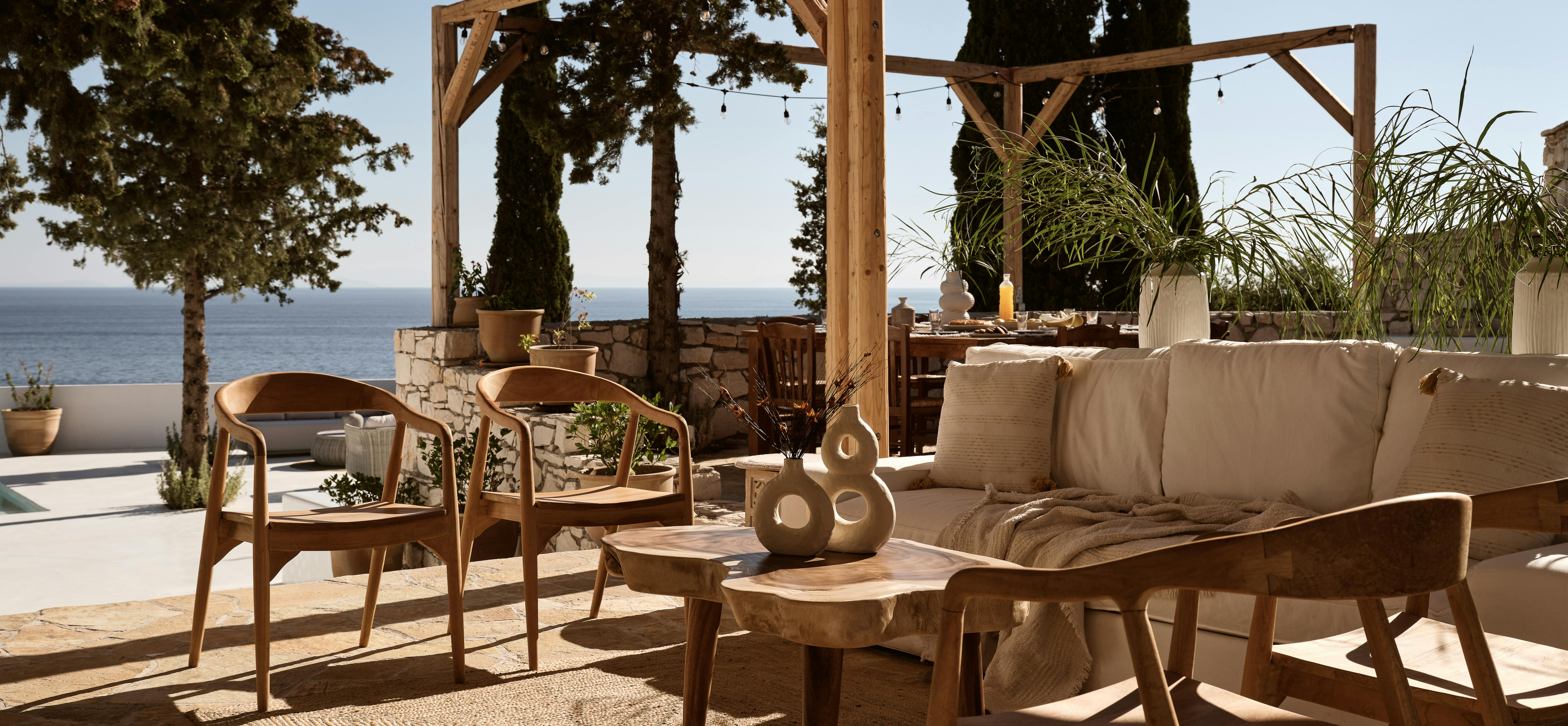 A covered outdoor lounge area with wooden furniture and neutral cushions opens to ocean views, with stone walls and string lighting overhead.