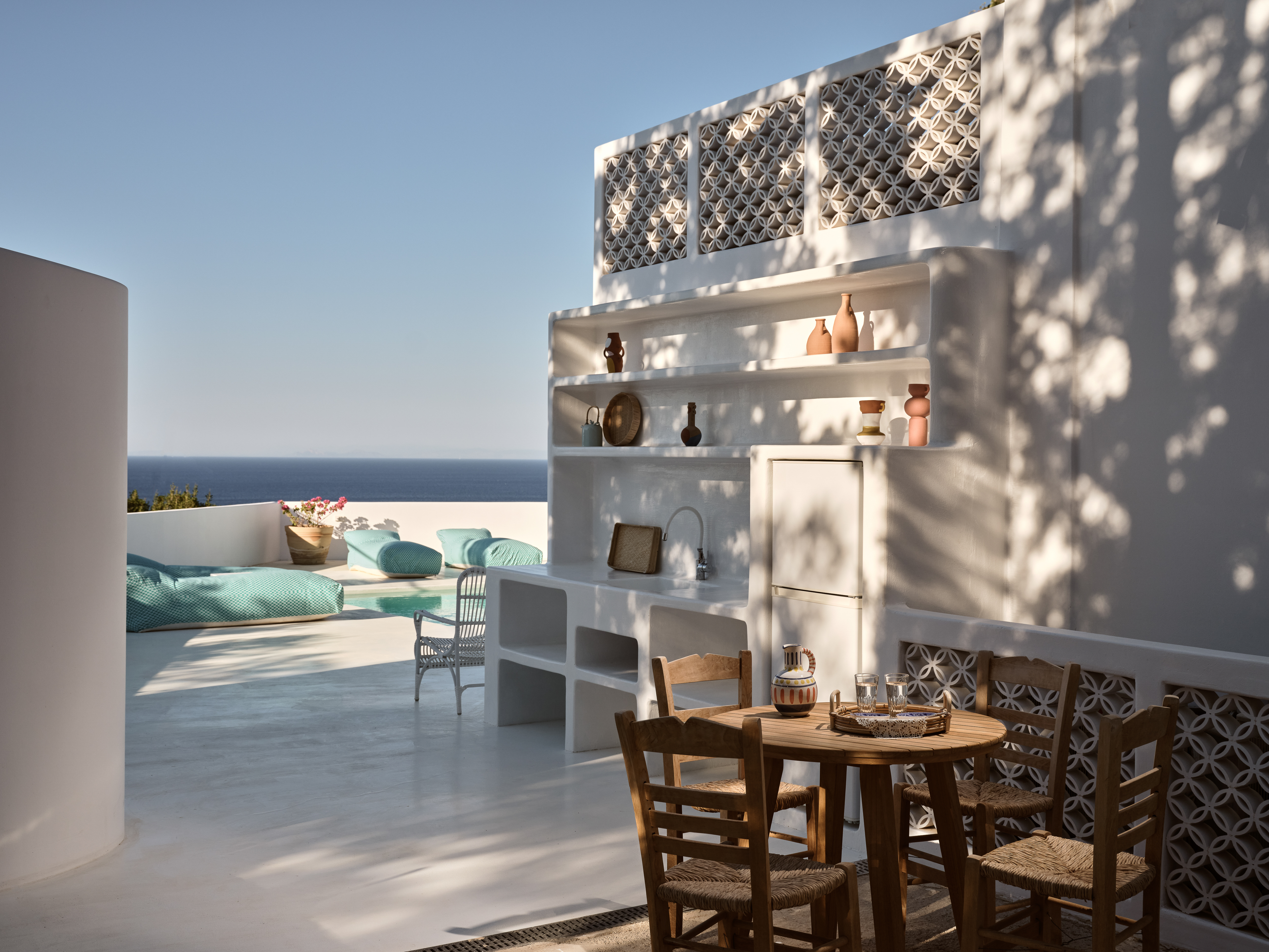 A white outdoor kitchen area with built-in shelving displays pottery and connects to the pool terrace, featuring decorative lattice screens and wooden dining furniture.