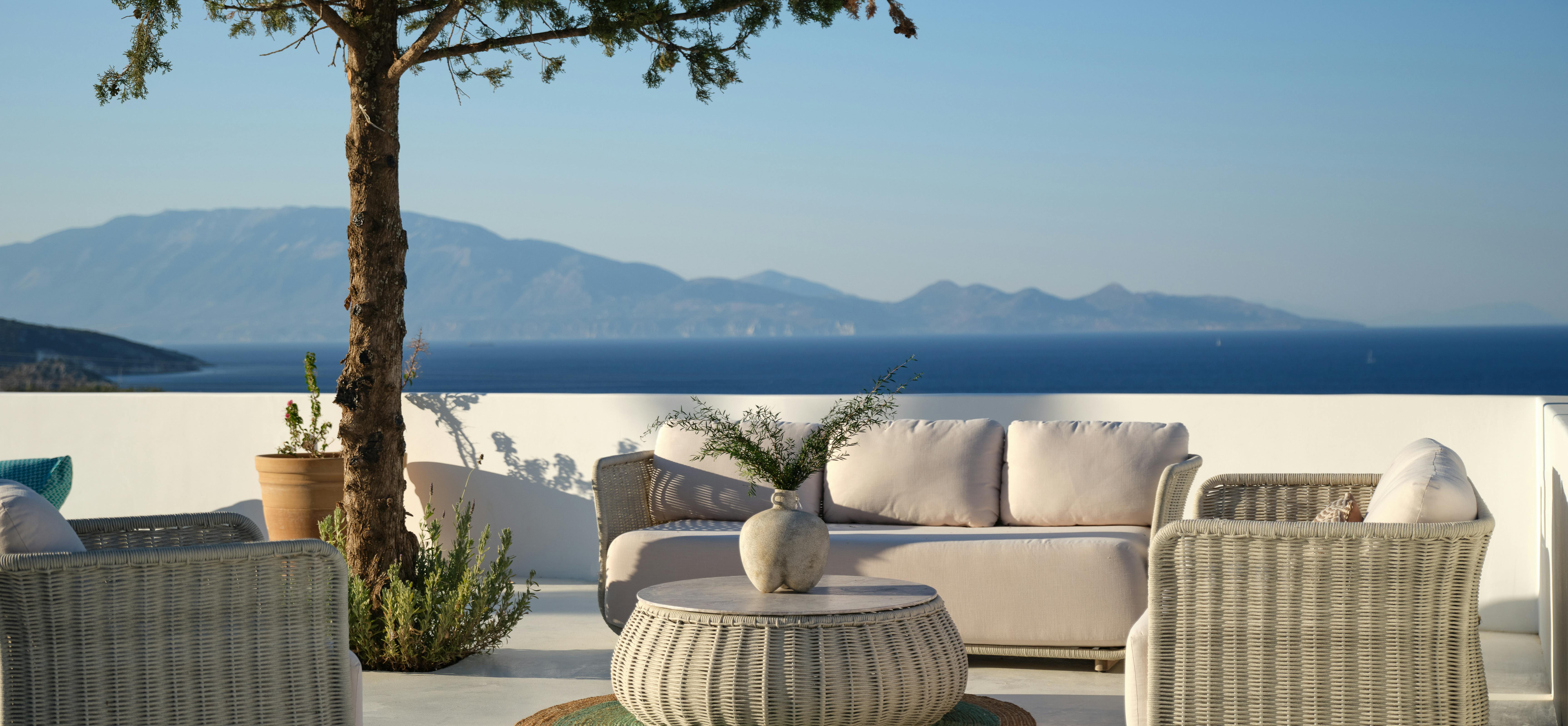 A minimalist white rooftop terrace features wicker furniture with cream cushions arranged around a low ottoman table beneath a lone tree, with mountain and ocean views beyond.