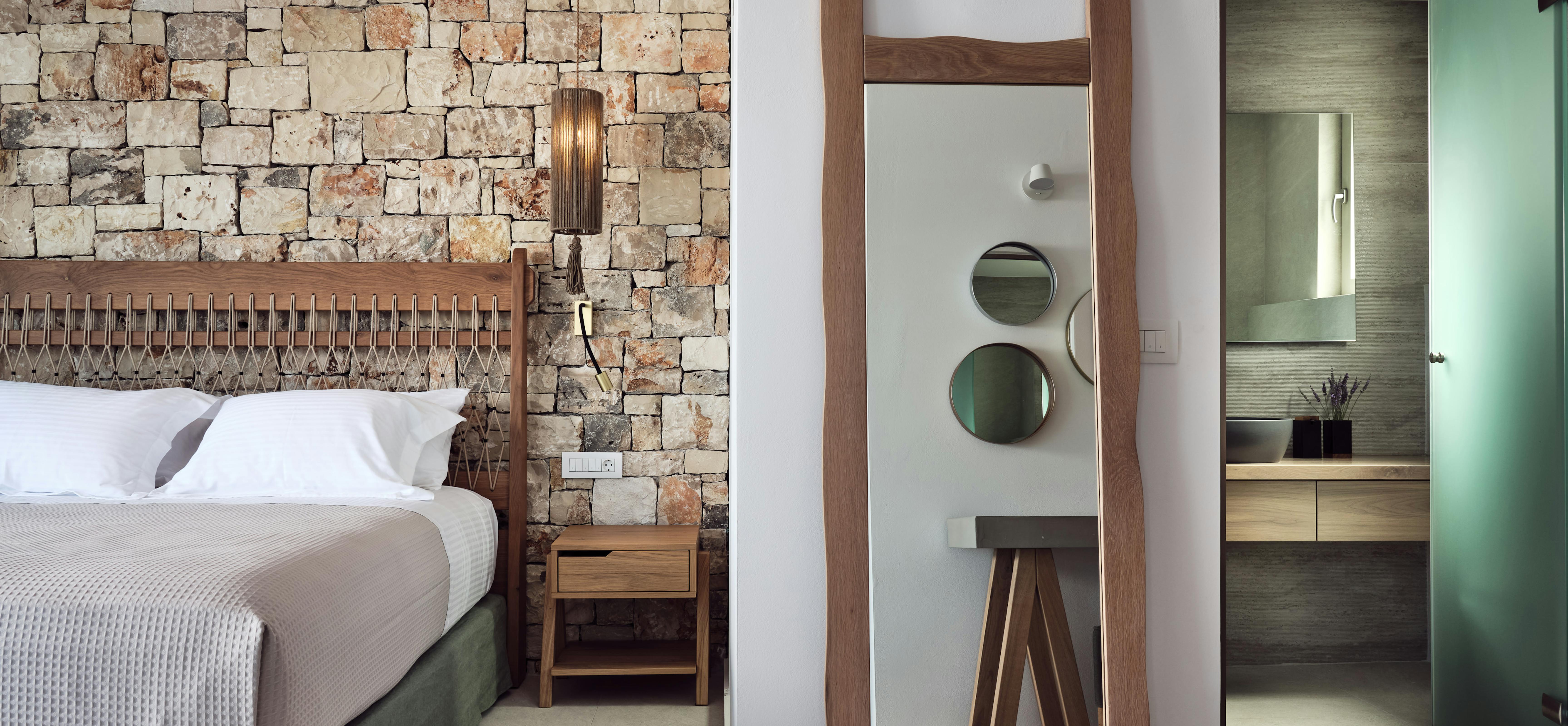A contemporary bedroom features a rustic stone accent wall, woven headboard, wooden nightstand, and tall mirror, with views into a mint-green accented ensuite bathroom.