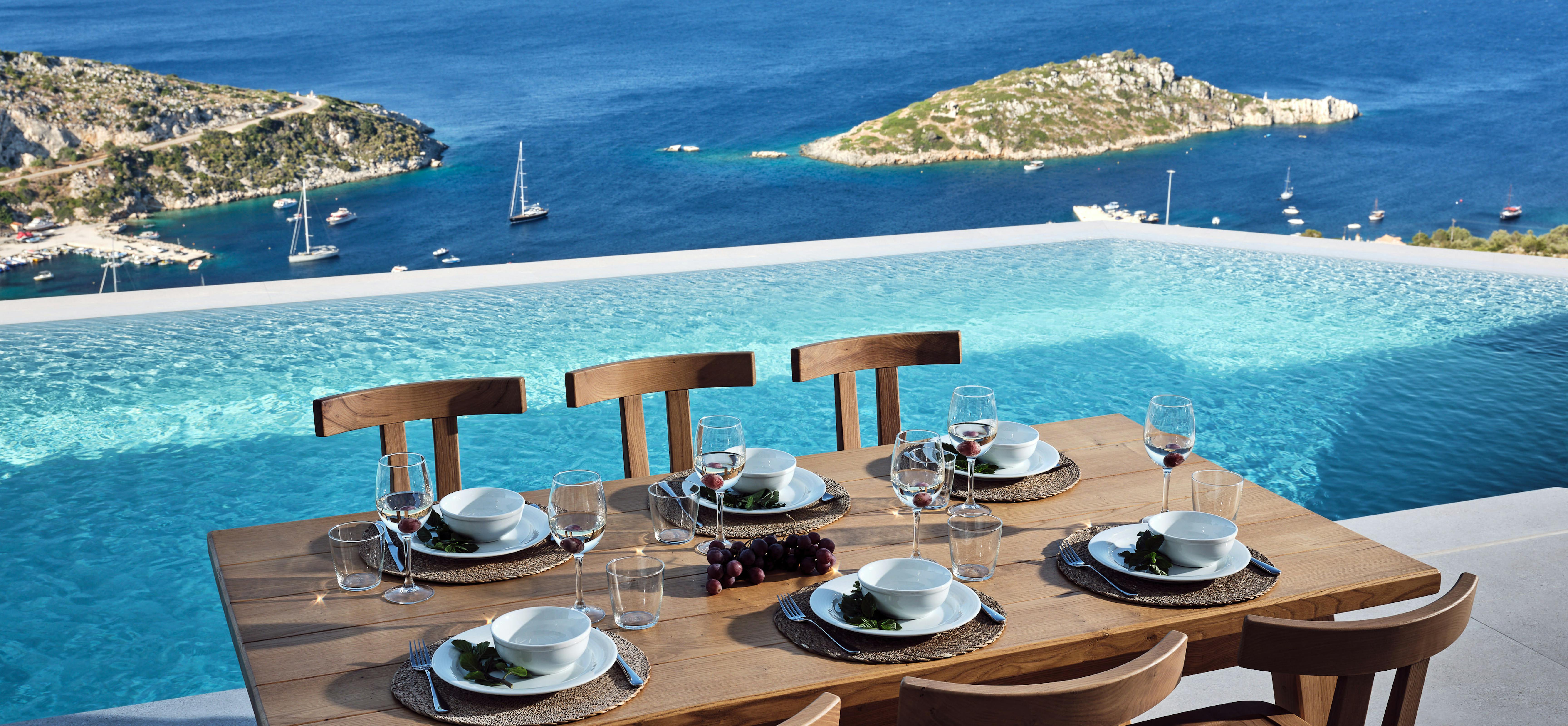 An elegant wooden dining table set for six with white plates, wine glasses, and woven placemats sits poolside, overlooking a bay dotted with sailboats and rocky islands.