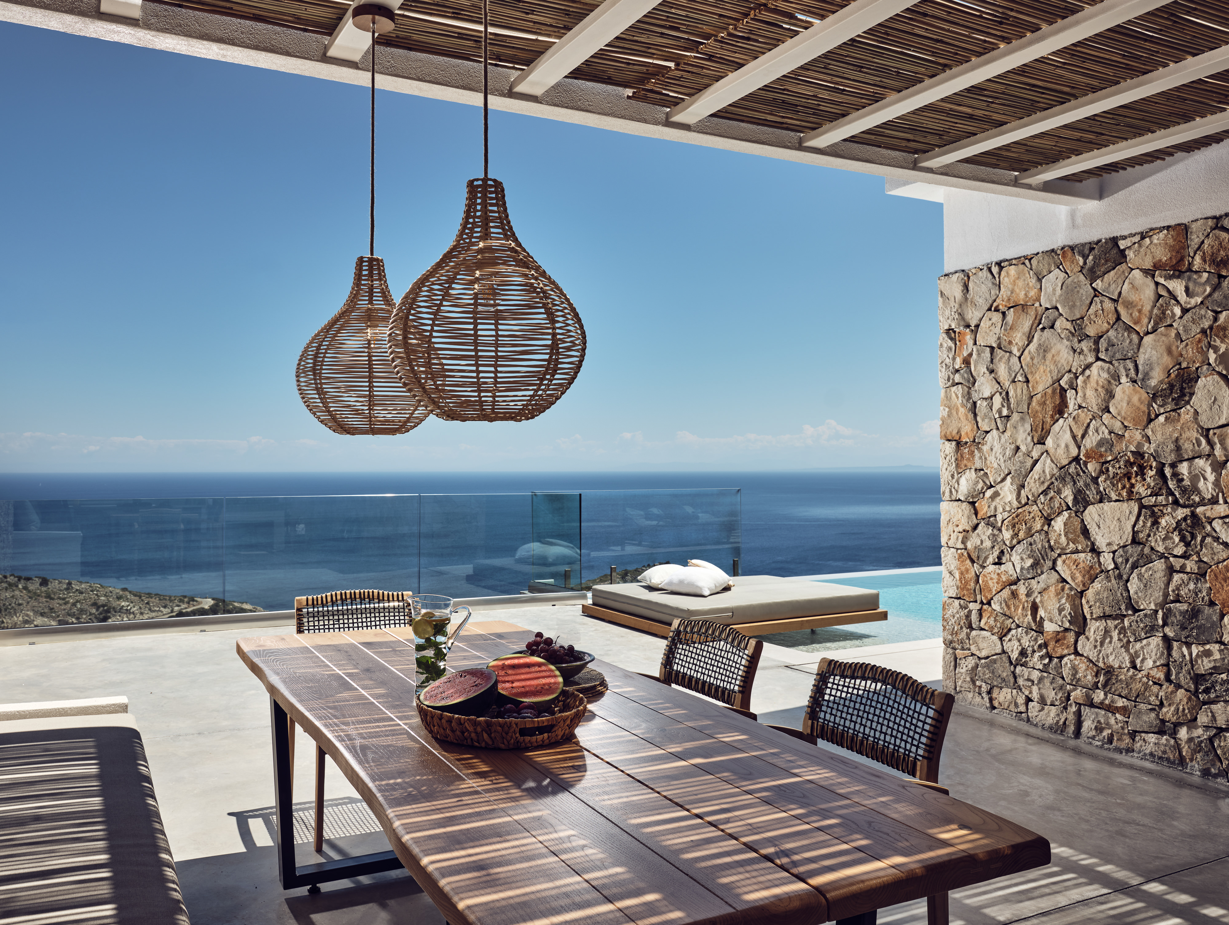 A covered dining terrace with wooden table and woven pendant lights frames views of an infinity pool, daybed, glass railings, and the sparkling sea beyond stone walls.