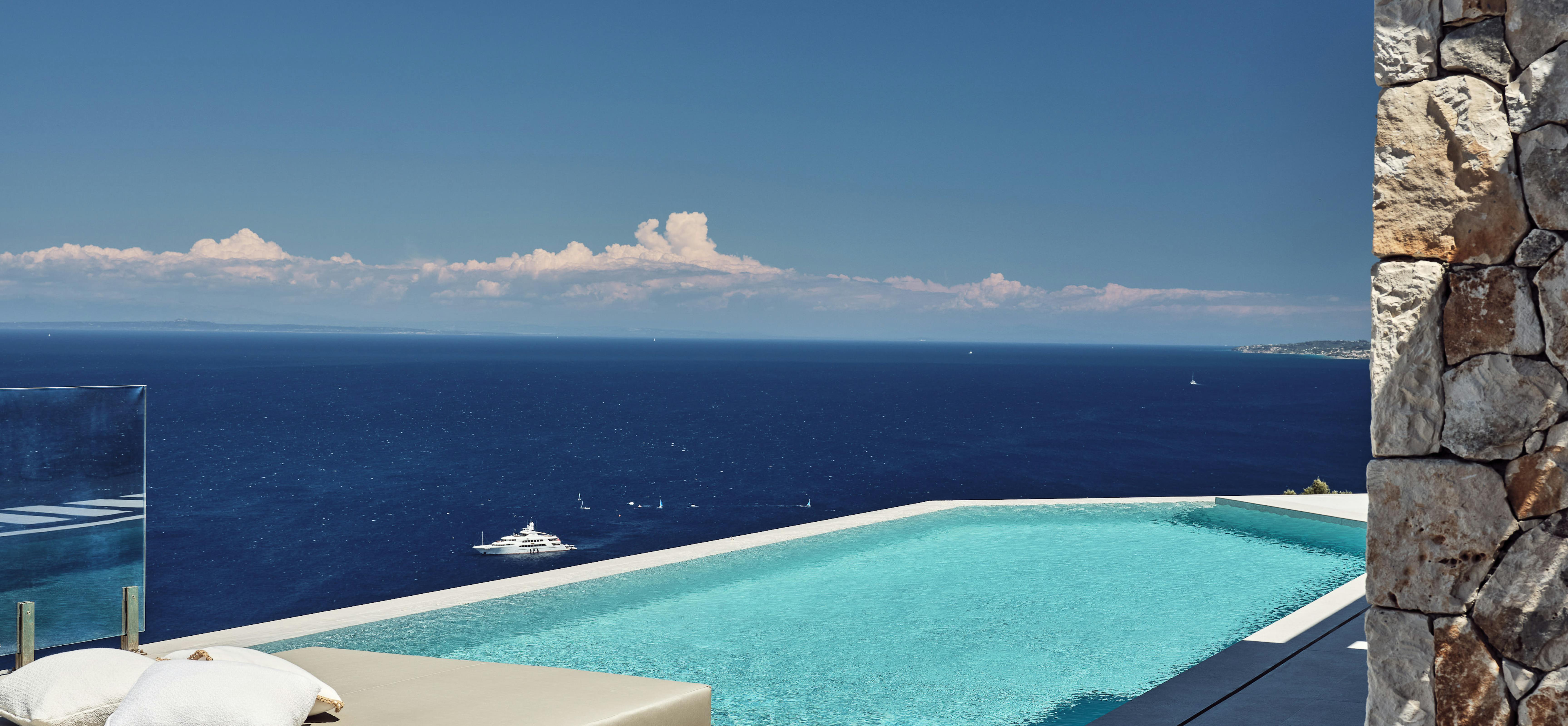A breathtaking infinity pool stretches toward the horizon where a yacht floats on deep blue water beneath white clouds, with a wooden daybed and stone wall framing the view.