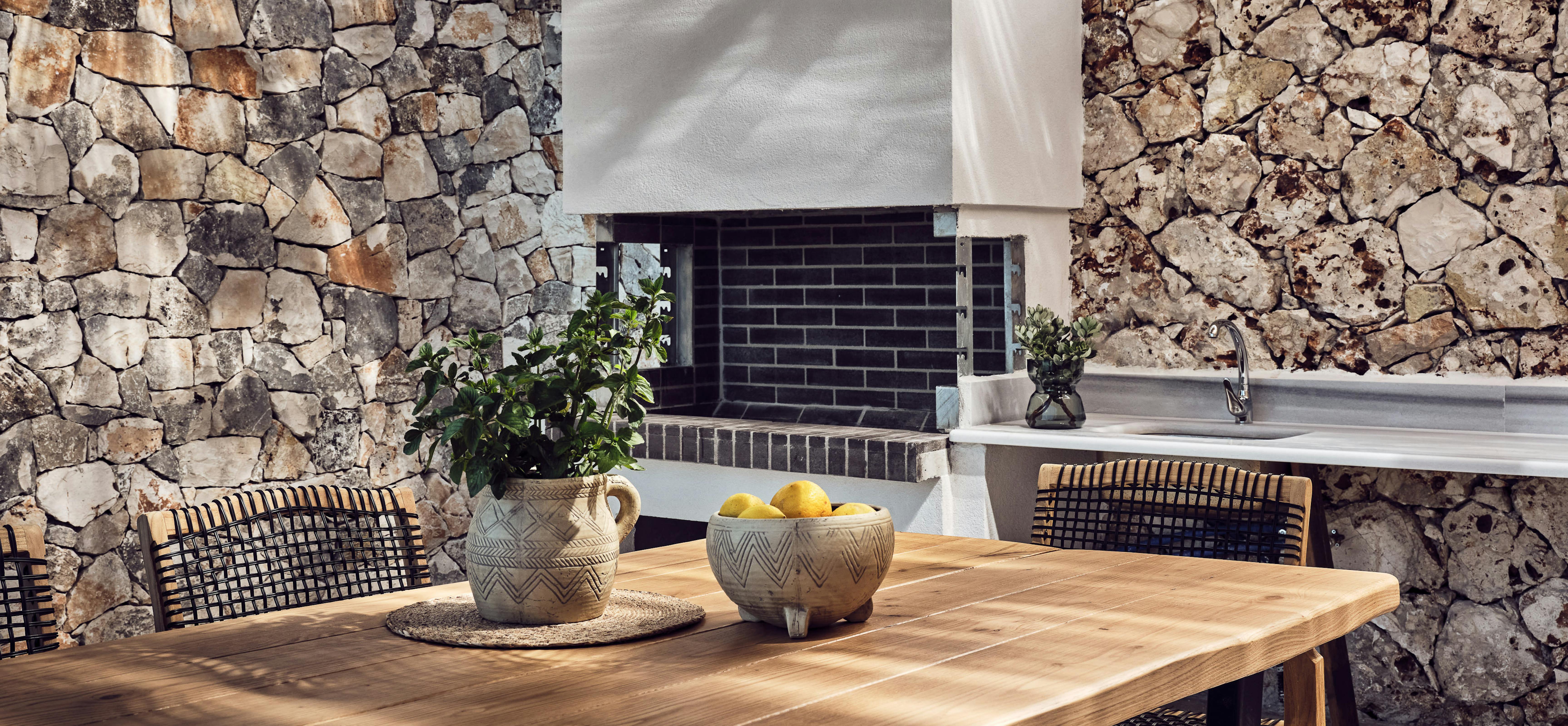 A rustic outdoor dining area with natural wood table, woven pendant lights, black woven dining chairs, ceramic bowls with lemons and greenery, flanked by textured stone walls and a built-in grill.