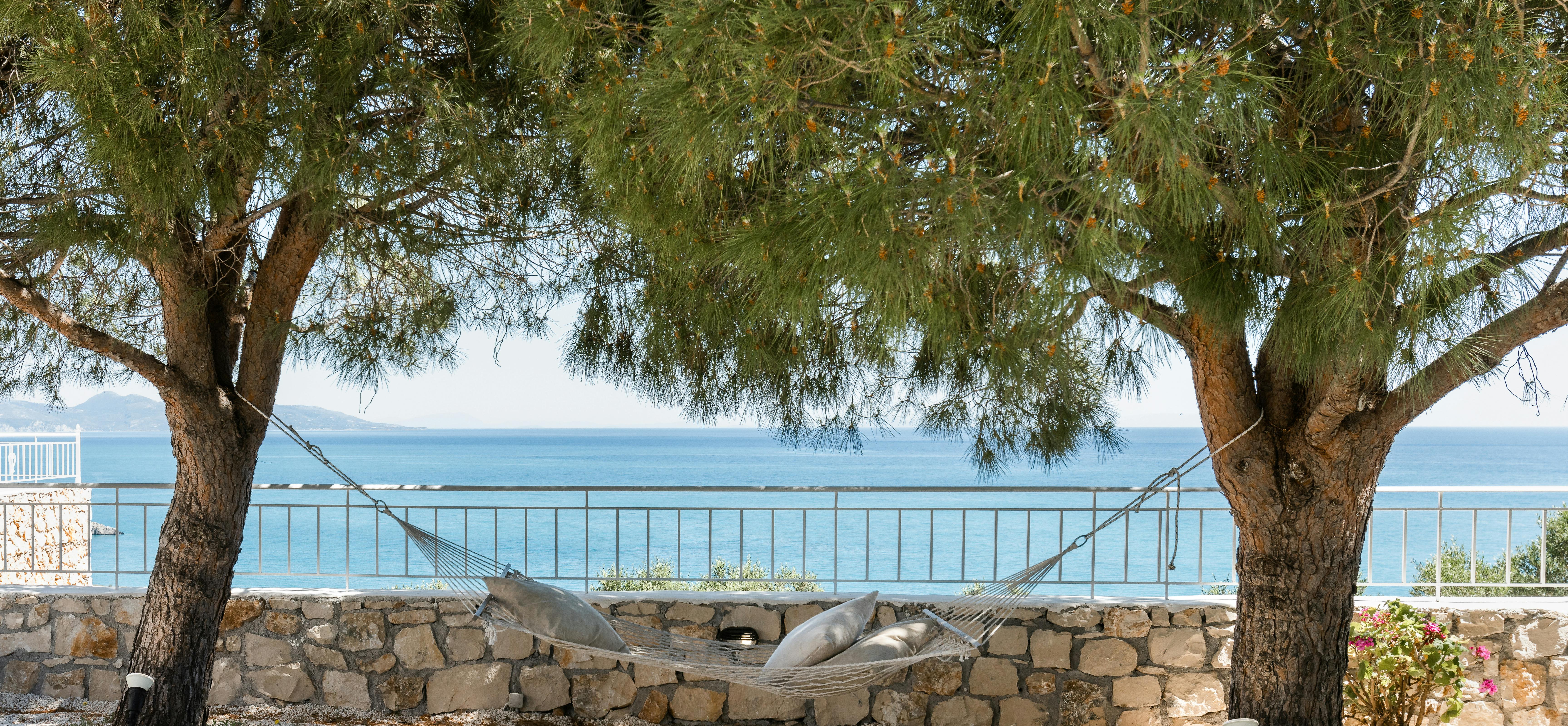 Hammock suspended between two pine trees overlooking the sea with stone wall and metal railing.